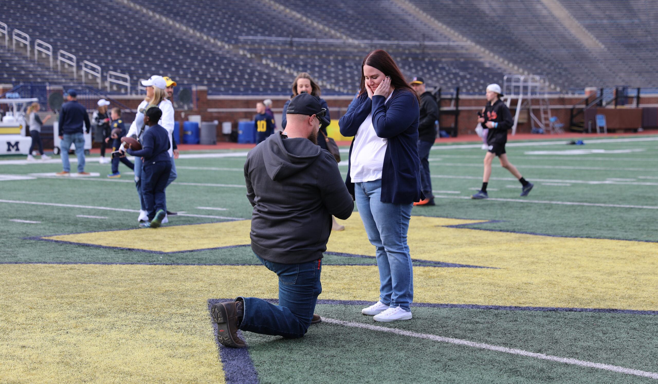 A man is on one knee on the Michigan Stadium football field and a woman has a surprised expression as she holds her hands against her face.