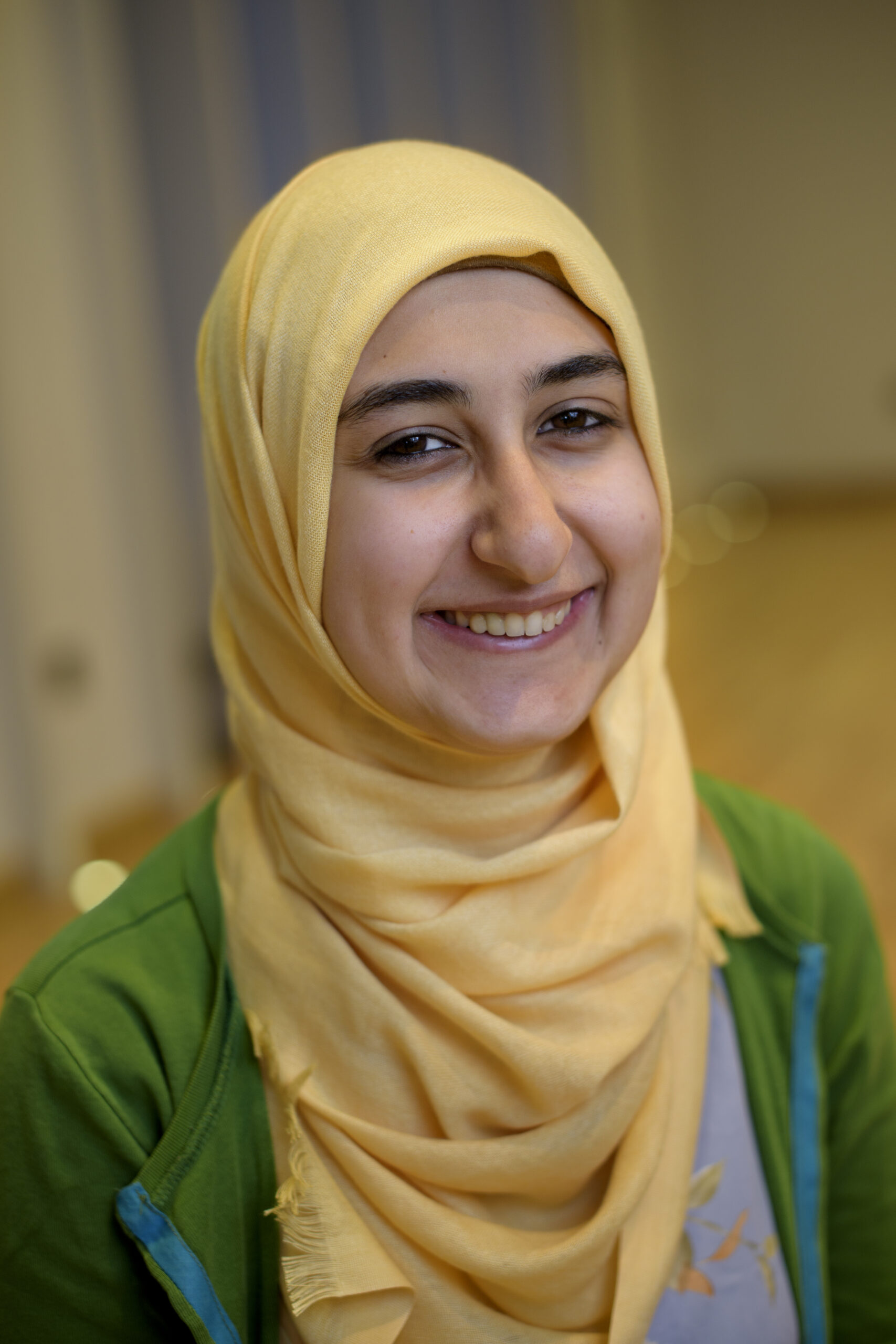A headshot of Jumanah Saadeh, who is wearing a green top and a yellow head wrap. 