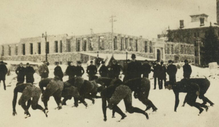 Black-and-white photo, tinged a pale yellow, of the Diag circa 1901 during winter. Most prominent in the photo is the group of practicing football athletes, lined up in a formation while a coach (possibly Fielding H. Yost) and other men (possibly other athletes or assistant coaches and staffers) look on. The ground is covered in snpw, and in the background is the mid-construction foundation of another University of Michigan building.