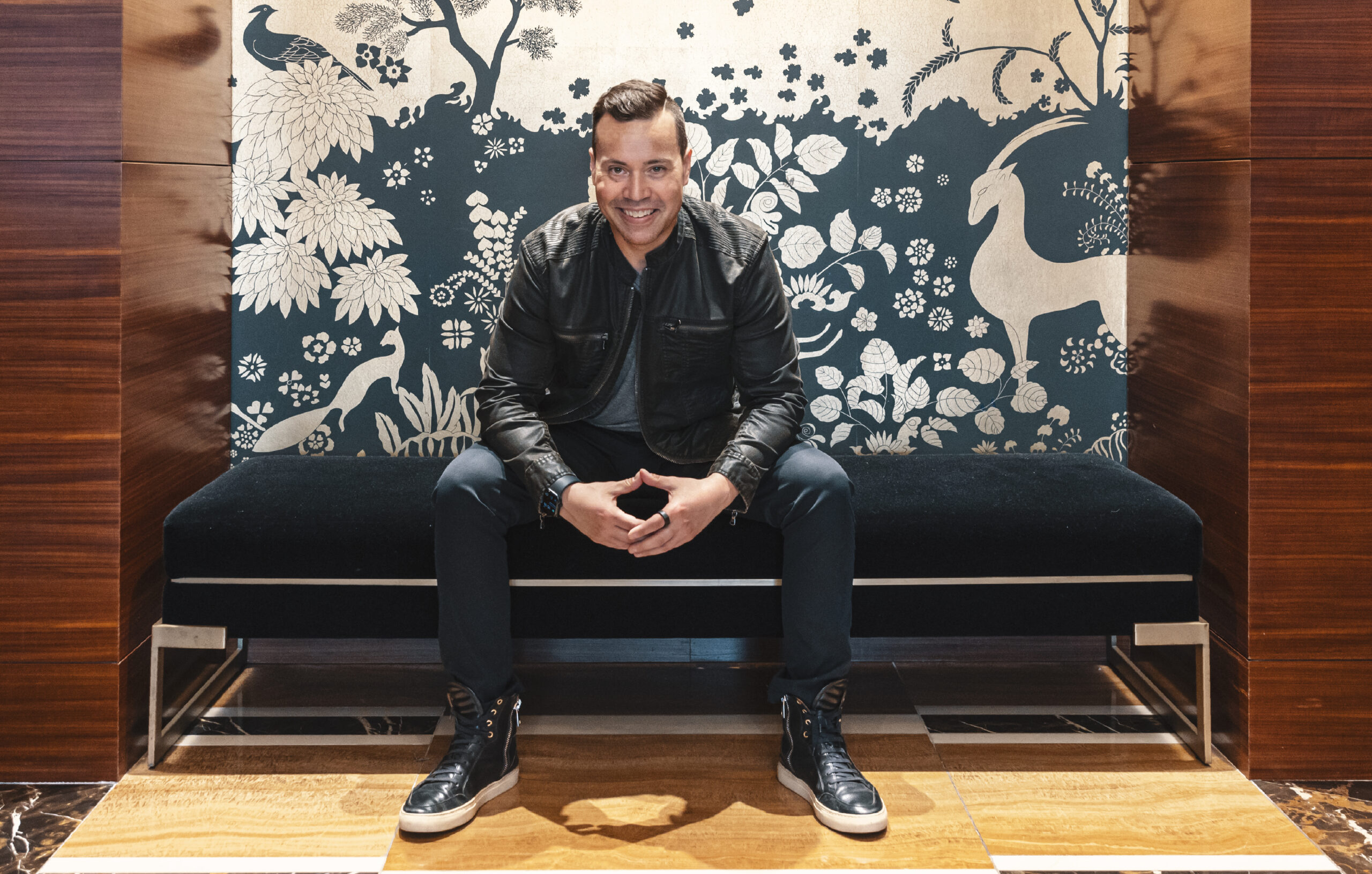 Jordan Harbinger, in a dark jacket, pants, and black shoes, poses on a bench with decorative wallpaper behind him.