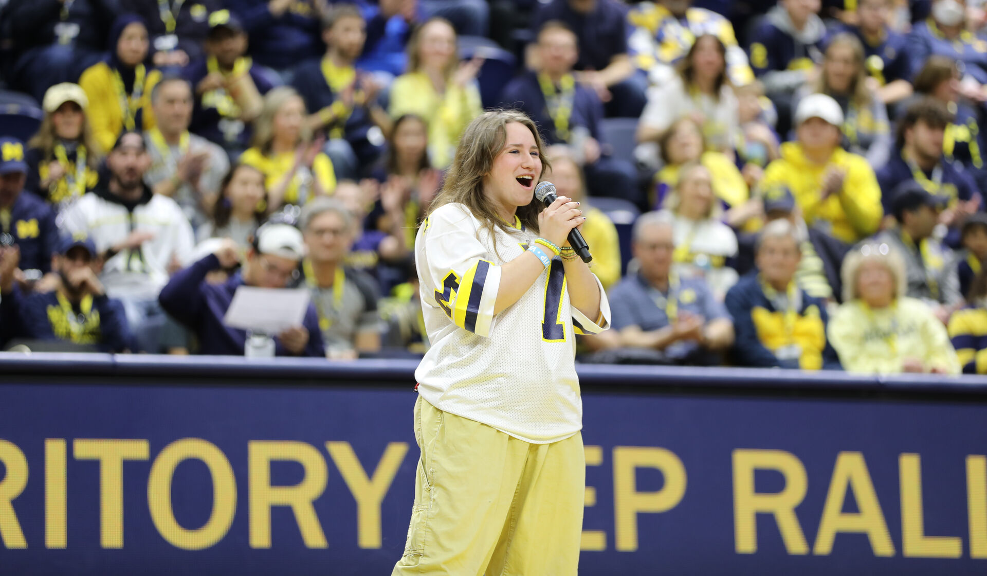 Daisy Sklar, in a white U-M jersey and khaki pants, speaks into a microphone with a maize and blue crowd behind her.