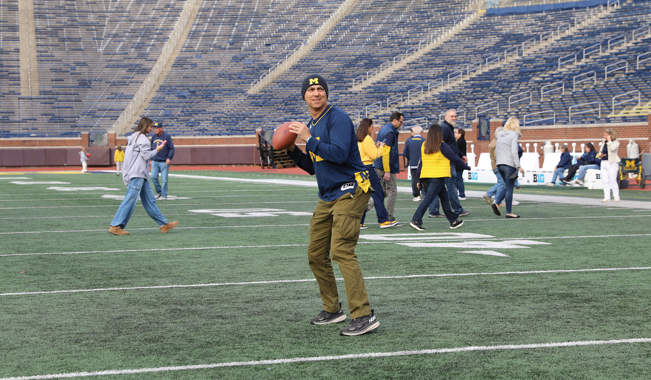 A person wearing Michigan gear readies to throw a football on the field of Michigan Stadium, surrounded by fellow attendees during a Big House Tour.