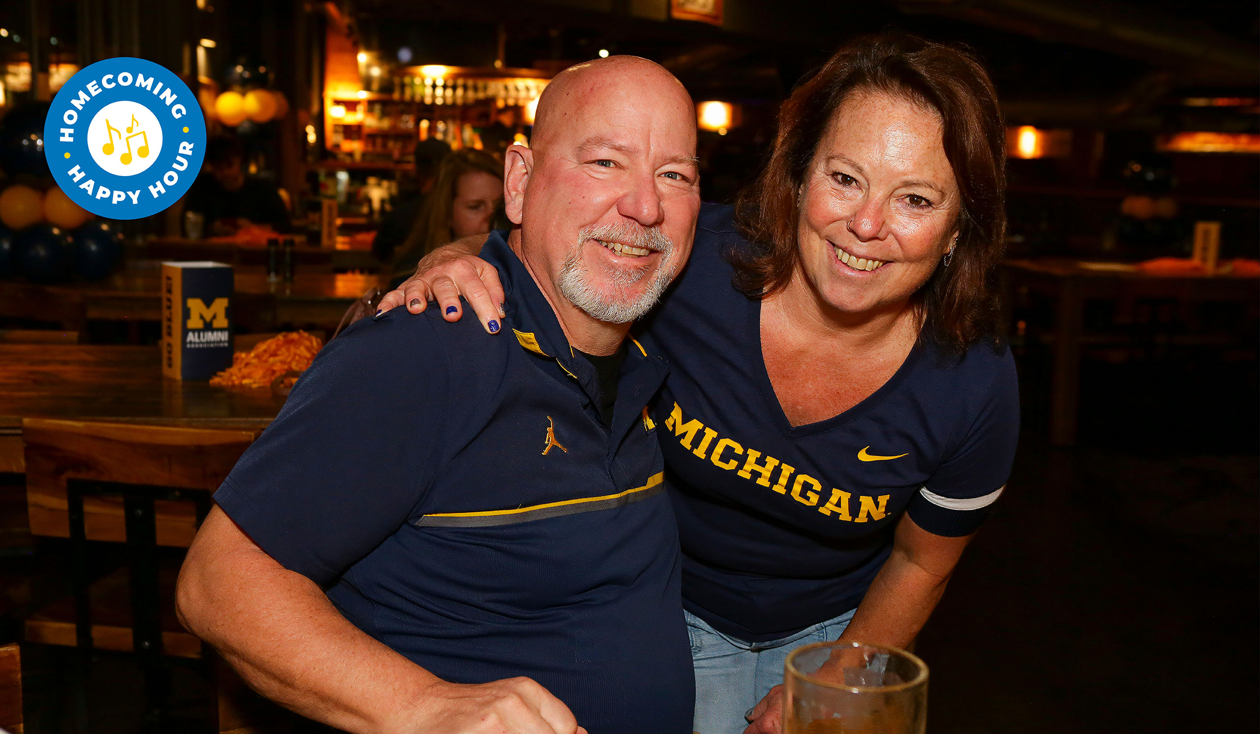 Two people in Michigan shirts pose together at a casual indoor event decorated with maize and blue. In the upper-left corner is an event icon for the Homecoming Happy Hour.
