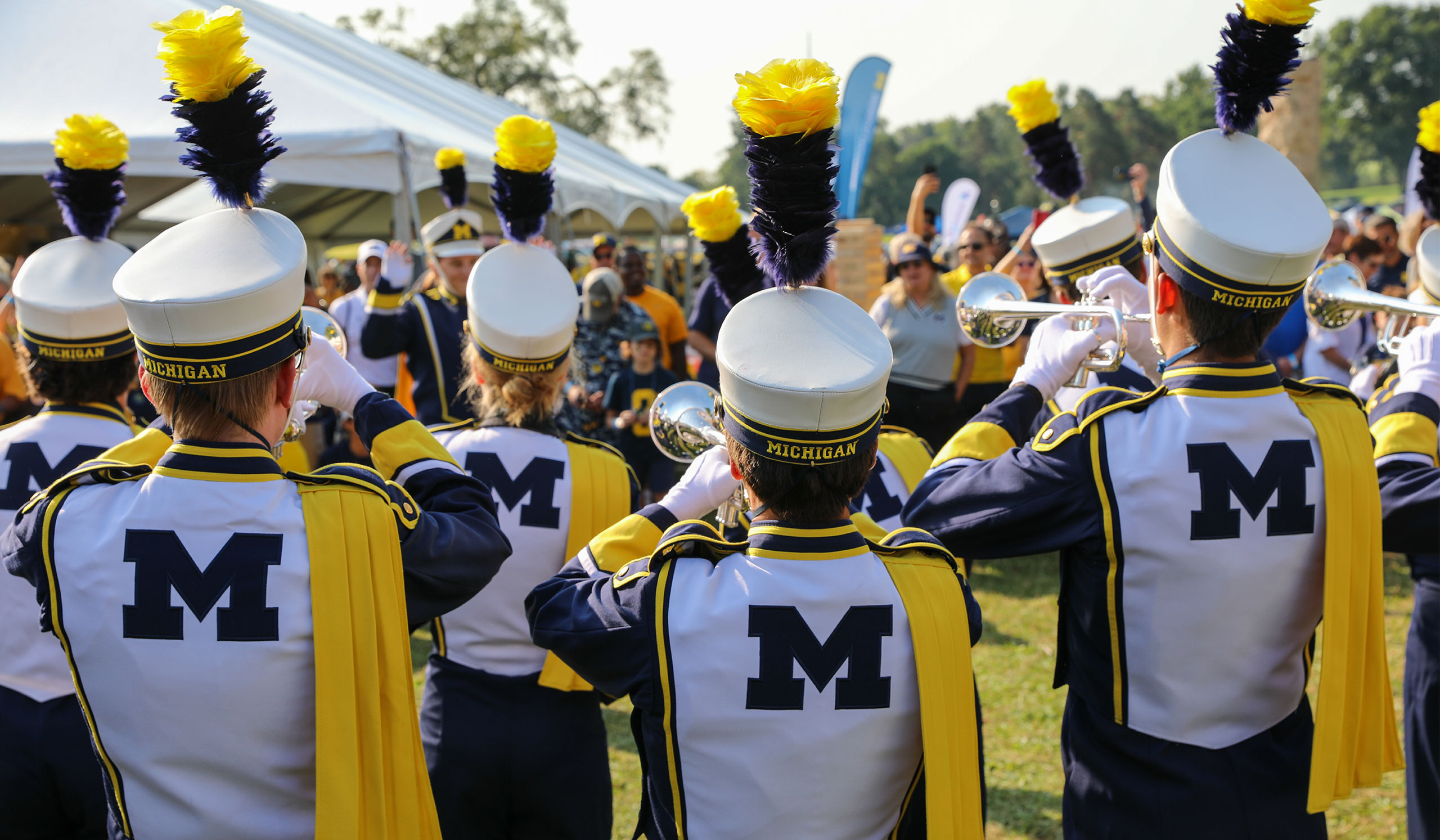 Michigan Marching Band members play at the 2024 Alumni Territory Homecoming Tailgate, with a tent and crowd in the background.