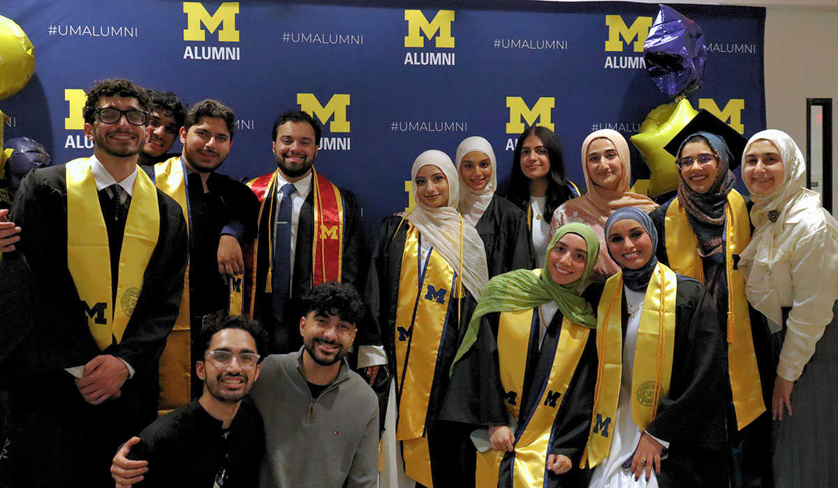 14 graduates pose before a University of Michigan alumni backdrop.