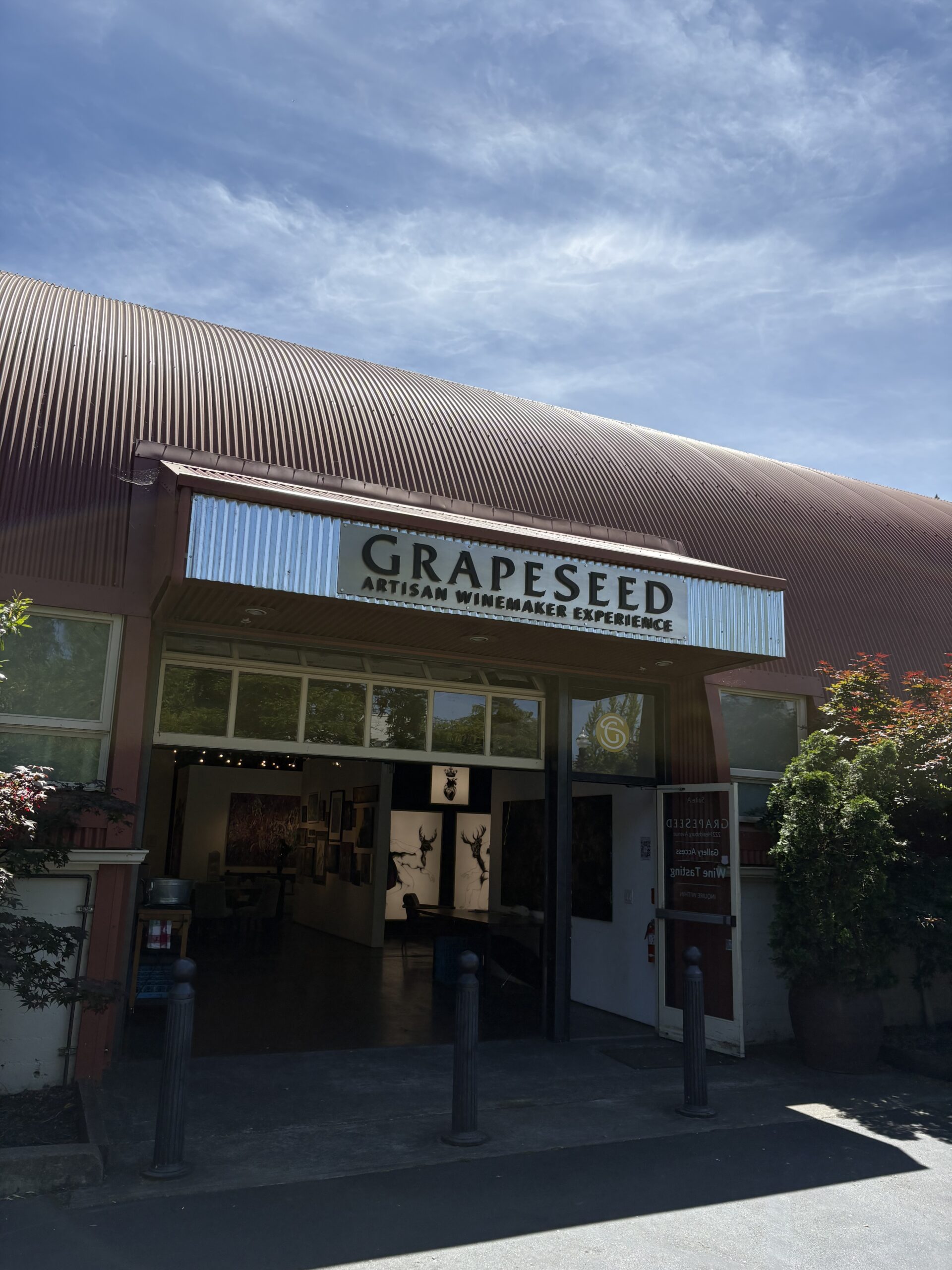An arched building with a corrugated metal roof has a sign above the entrance reading GRAPESEED Artisan Winemaker Experience. Once host to a Sacramento Club past event, its open doorway reveals artwork inside, framed by trees under a bright blue sky.