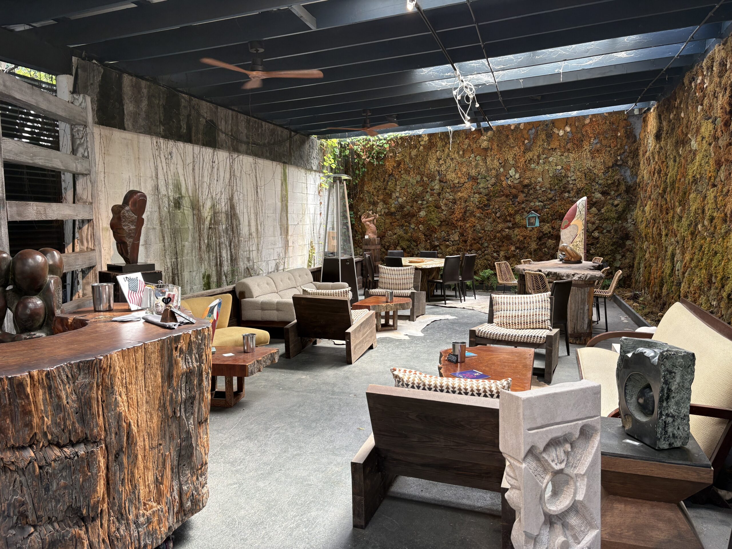 Covered patio at Sacramento Club with wooden furniture, sculptures, and abstract art. High ceiling with fans, stone and moss-covered walls, and artistic decor create a perfect spot for event photos under natural light.