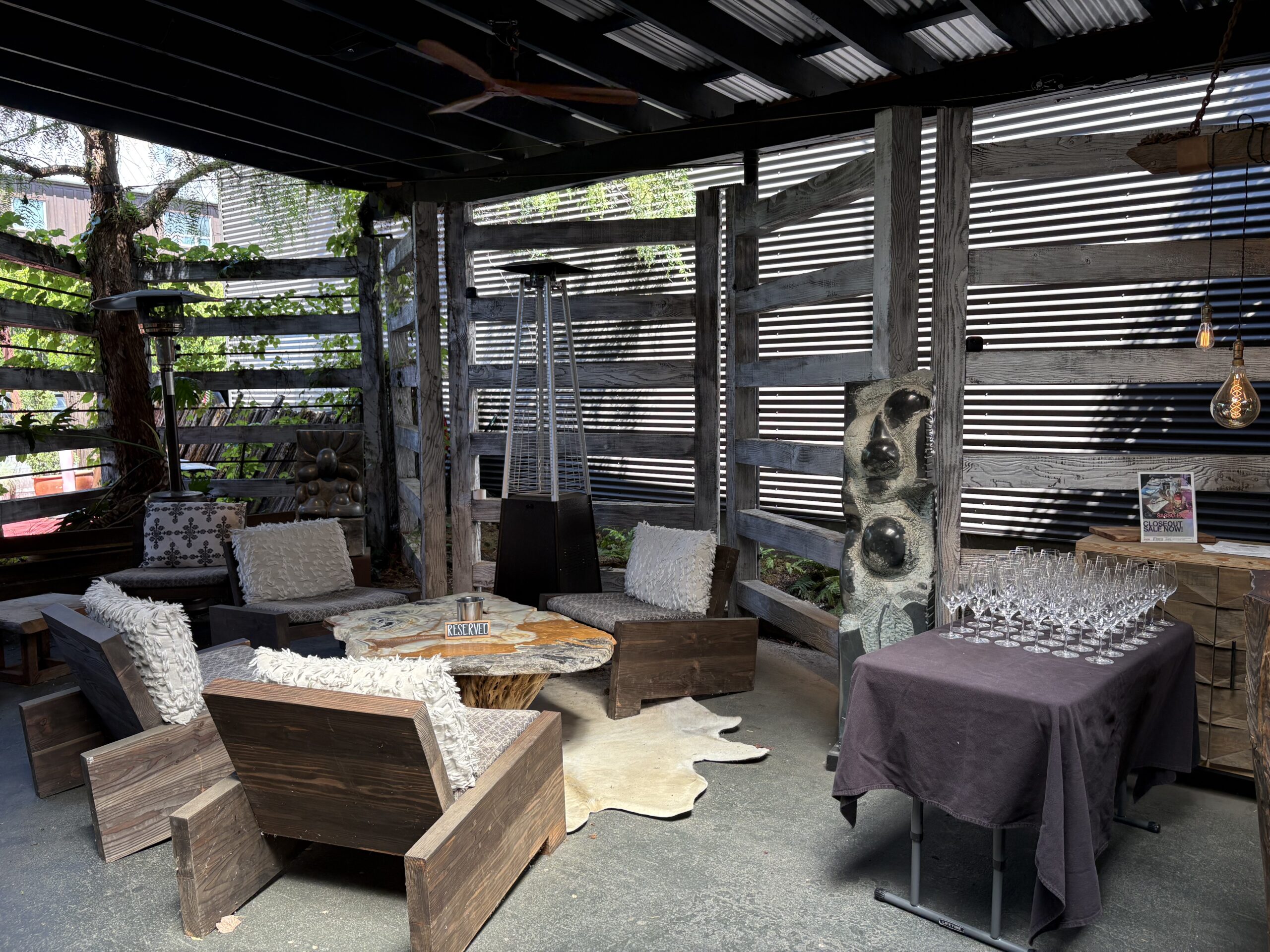 A modern outdoor lounge at Sacramento Club features wooden chairs, textured cushions, a cowhide rug, and sculptural art under a covered roof—perfect for Event Photos and recalling memorable Past Events amid slatted walls and lush greenery.