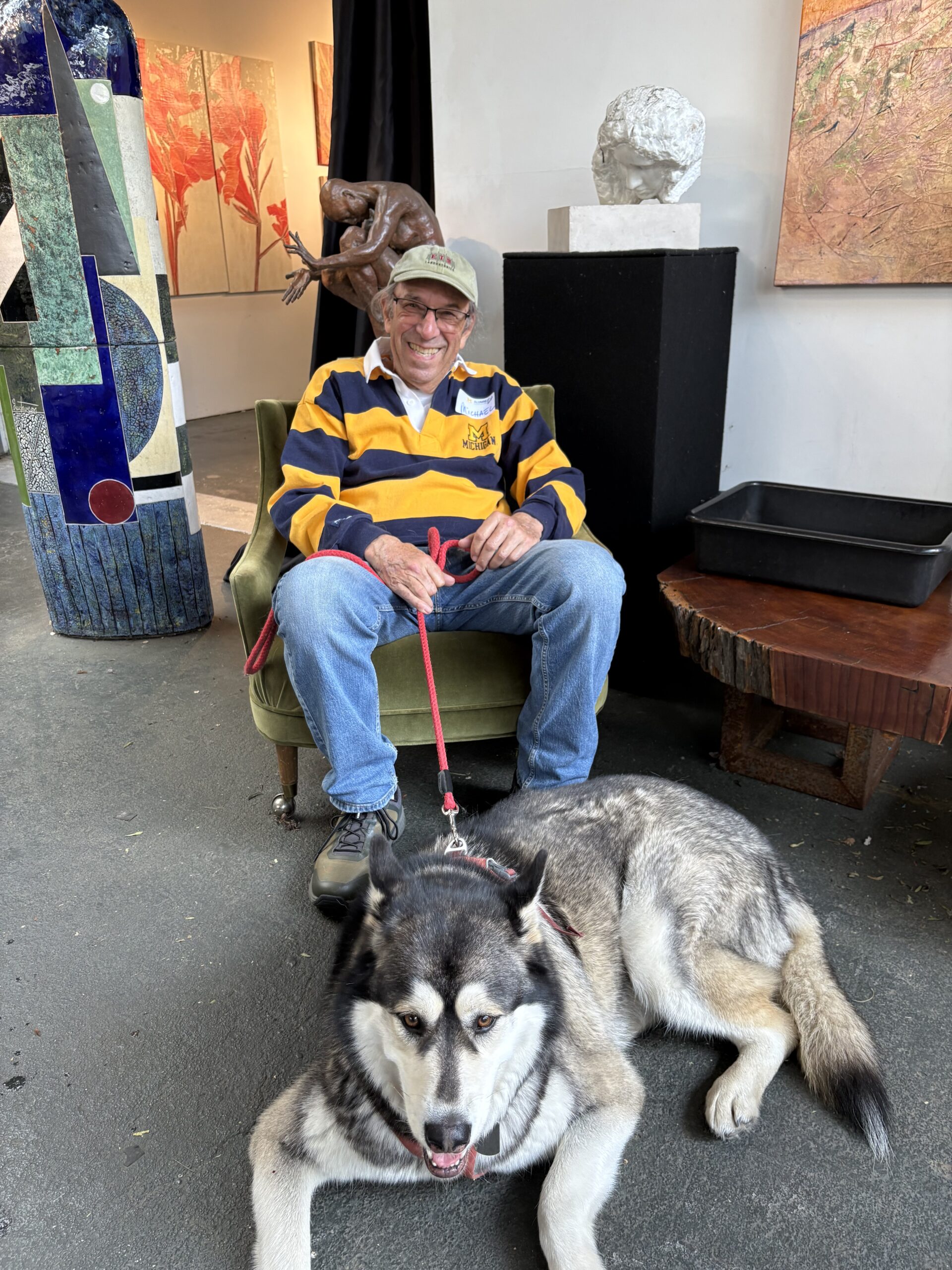 A man in a yellow and navy striped rugby shirt and cap sits smiling in a green chair, holding a red leash attached to a large, gray and white dog lying on the floor at an art club event in Sacramento.