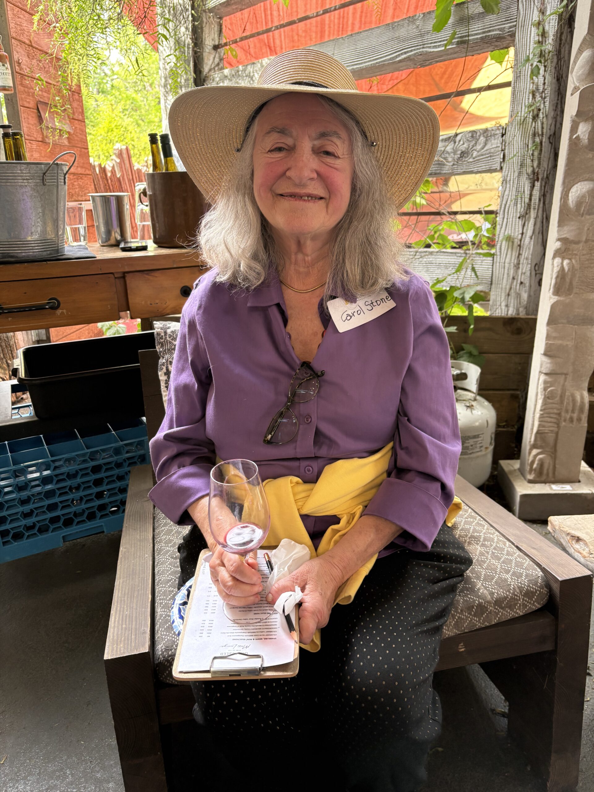 An older woman with long gray hair, wearing a wide-brimmed straw hat, purple shirt, and yellow sweater, smiles while holding a wine glass and pen. Seated outdoors in Sacramento, she enjoys the club event photos with a clipboard on her lap.