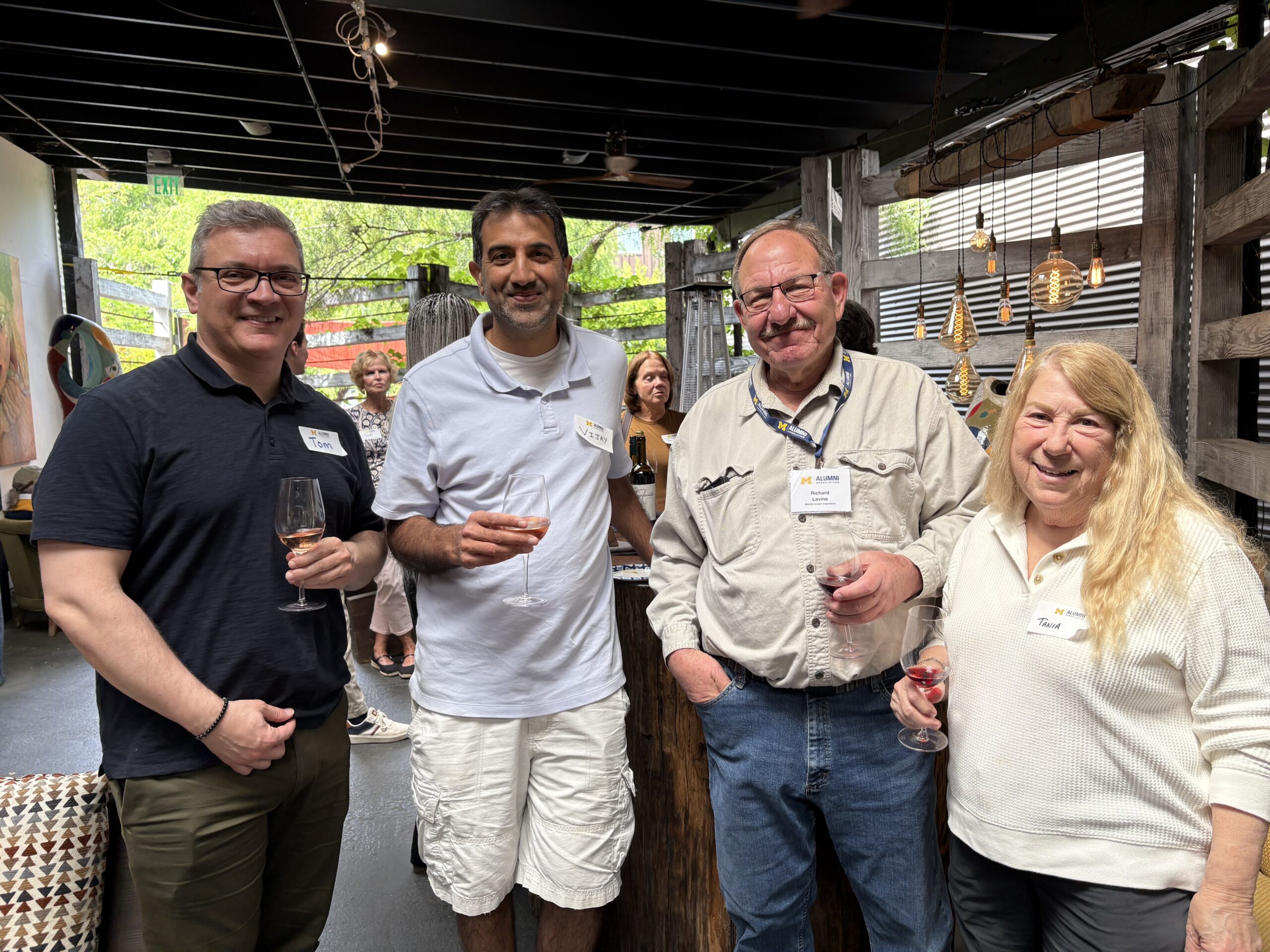 Four adults stand together indoors at a Sacramento Club event, each holding a drink and wearing name tags. The warmly lit setting features rustic decor, hanging lights, and other guests mingling—perfect for memorable event photos.