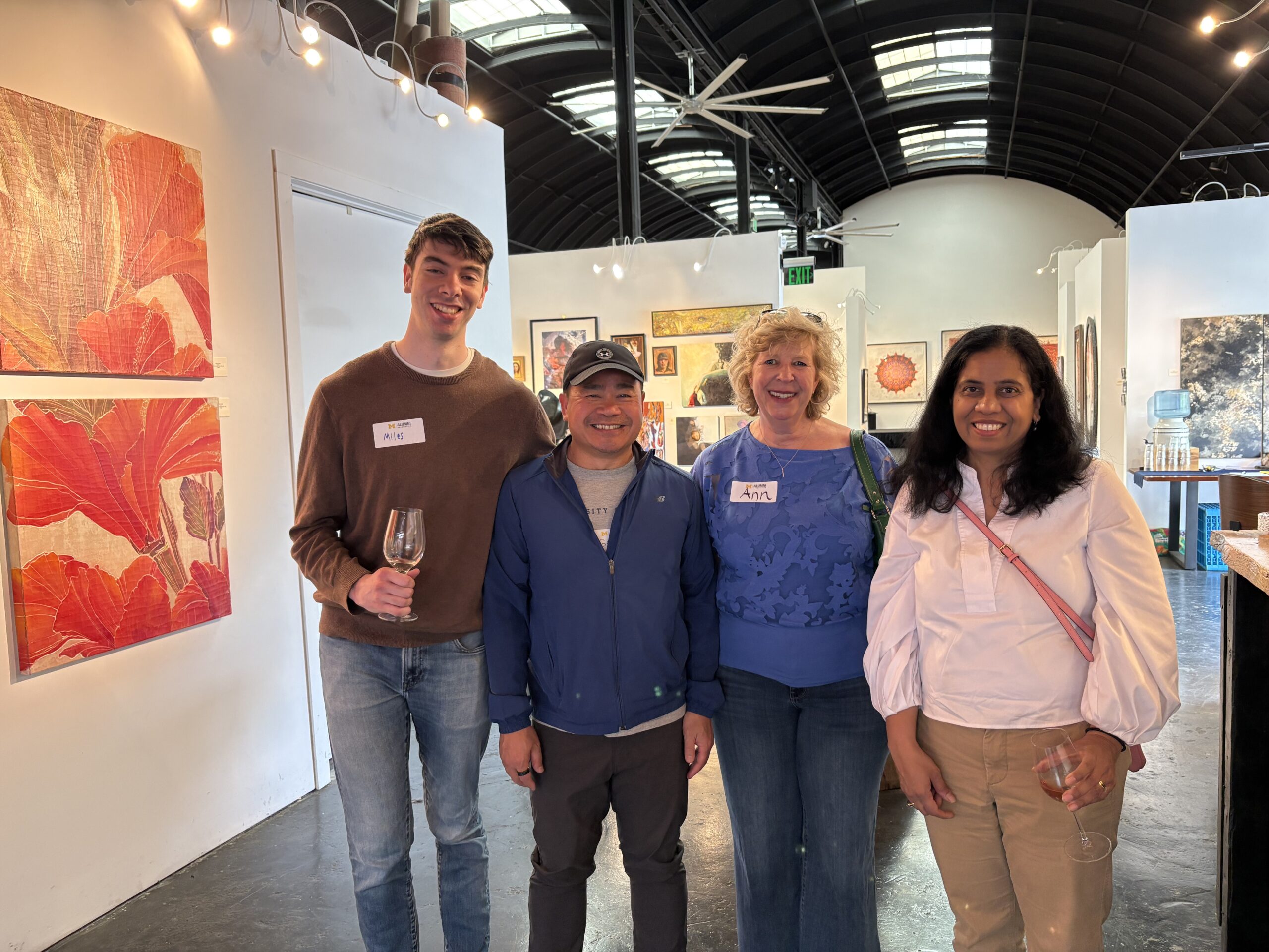 Four people stand and smile inside an art gallery with modern paintings on the walls. Two hold wine glasses. All wear name tags, and the bright space features arched ceilings—perfect for Sacramento Event Photos or Sacramento Club gatherings.