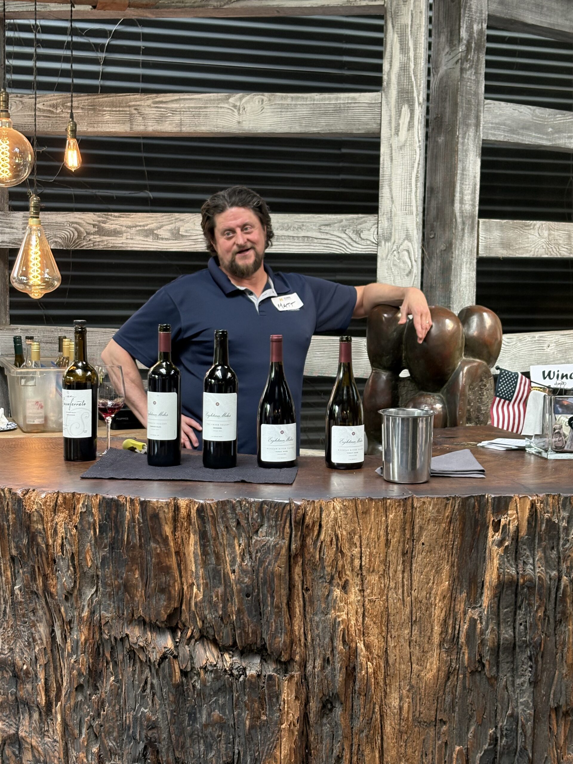 A man with wavy hair and a beard stands behind a rustic wooden bar at a Sacramento Club, five bottles of wine displayed. Vintage-style lights hang above, with an American flag and an ice bucket on the bar—perfect for event photos.