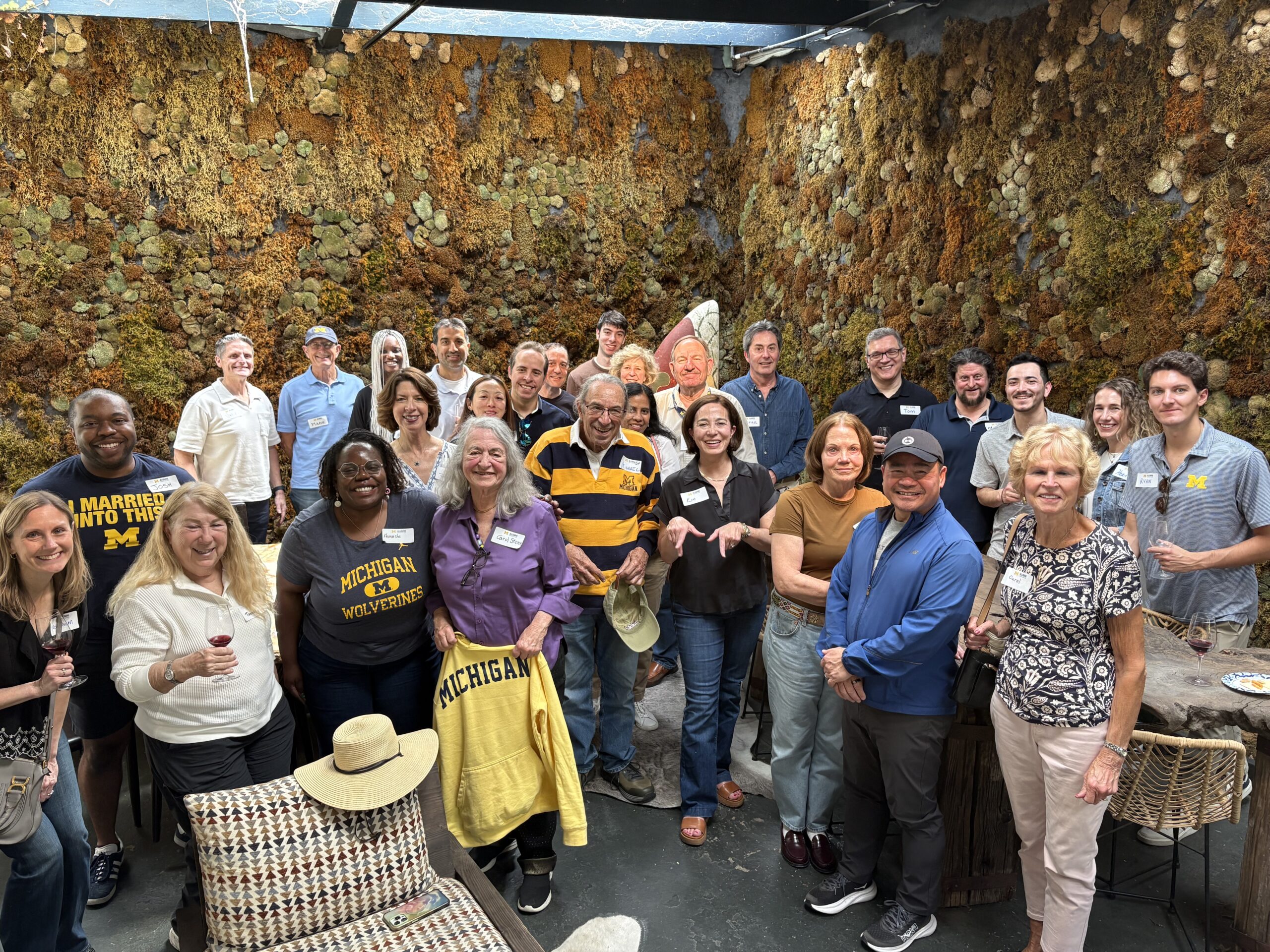 A group of adults, some in University of Michigan apparel, smile at the camera indoors. The Sacramento Club’s past event photos show them gathered by a plant-covered wall, holding drinks and enjoying a lively social gathering.