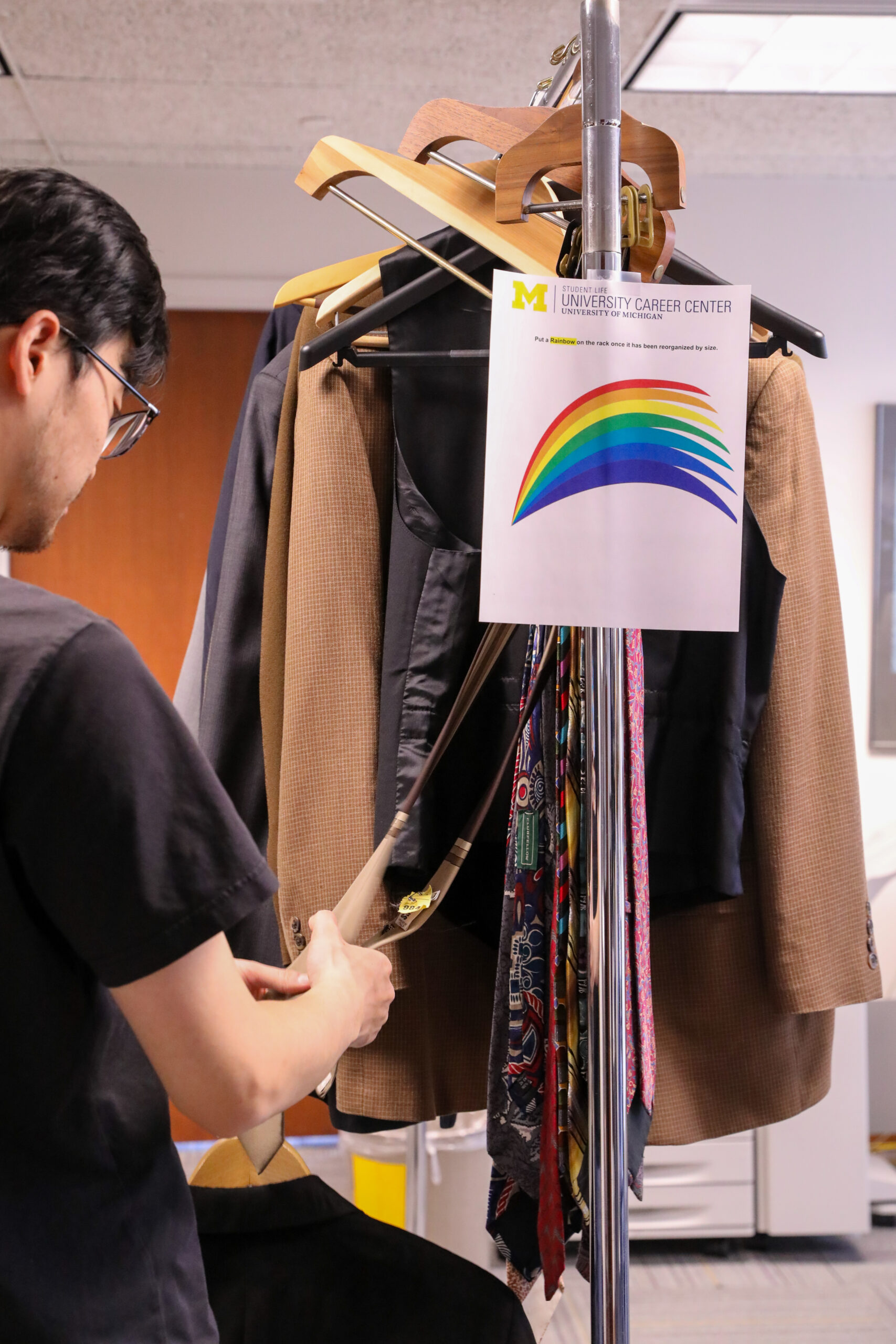 A male student sorts through ties on a clothing rack. The rack has a rainbow sign indicating the rack has been reorganized. 