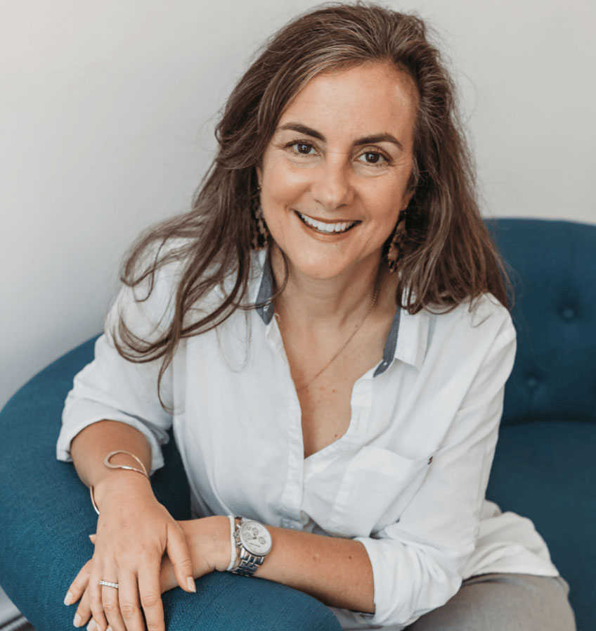 Hallie Crawford, with long brown hair and a white shirt, sits on a blue chair, smiling at the camera.