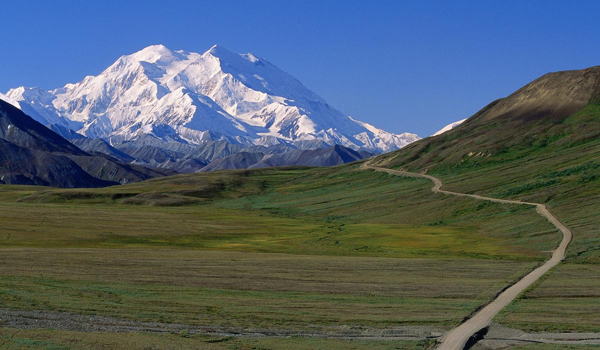 Alaska Untamed Wilderness Snowy mountain caps are in the background, while green mountains are in the foreground with a brown path connecting the two.