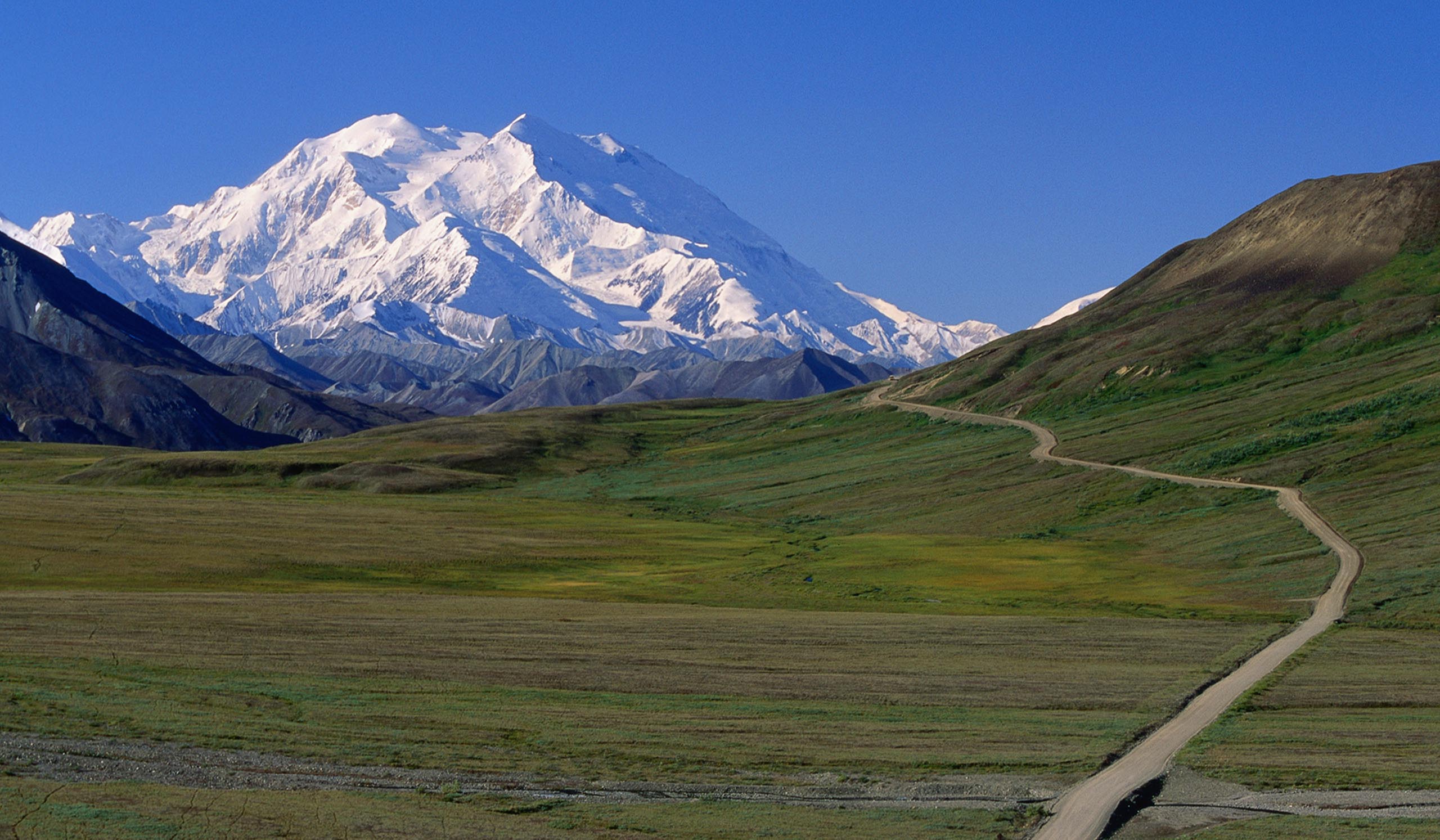 Snowy mountain caps are in the background, while green mountains are in the foreground with a brown path connecting the two.