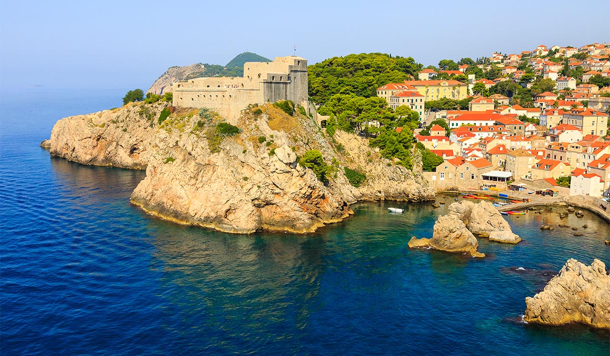 A coastal town perched on rocky cliffs, featuring a large stone fortress overlooking clear blue water. Below, a small harbor sits beside tightly clustered buildings with red-tiled roofs, surrounded by lush green trees and rugged shoreline.