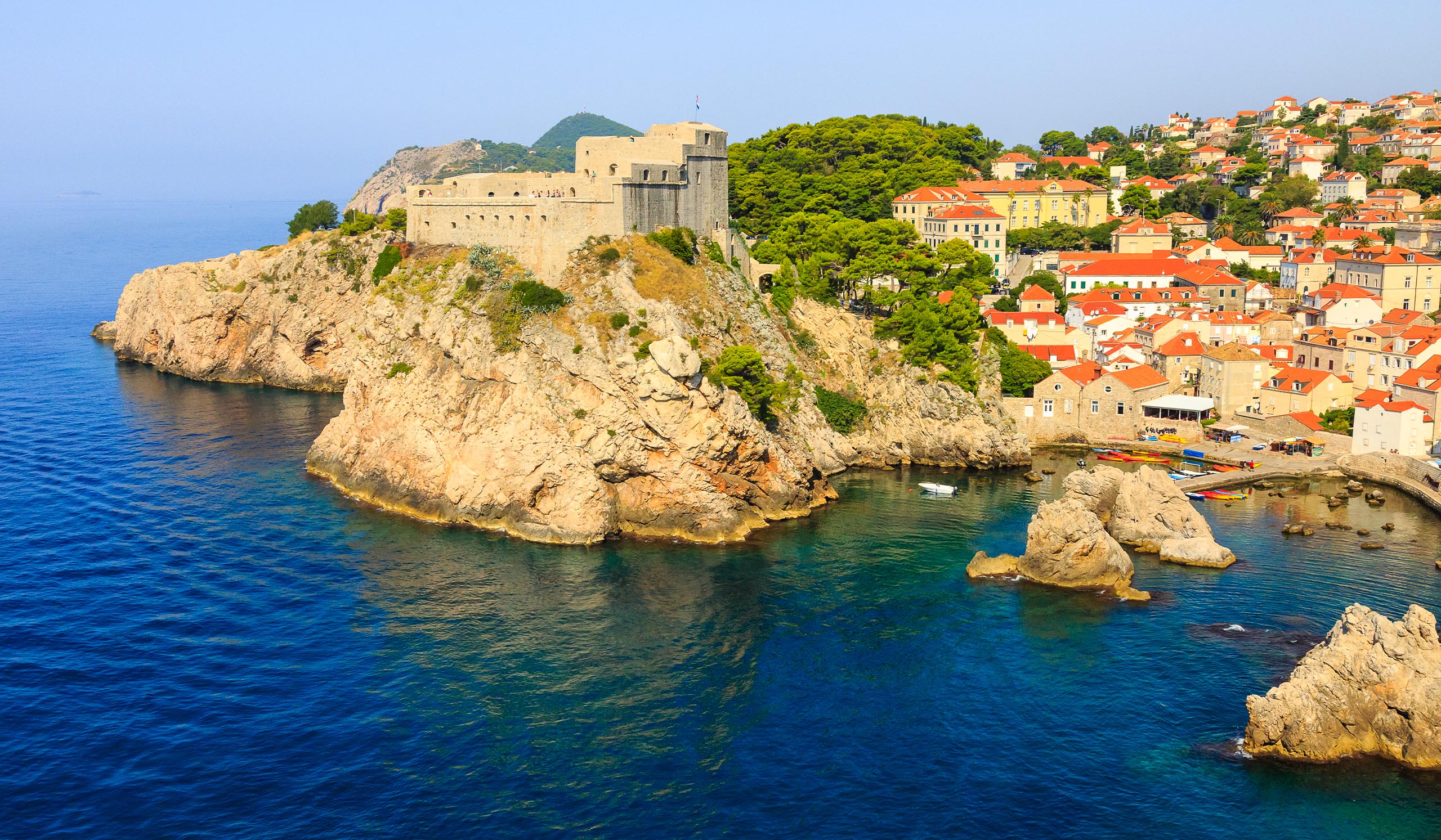 A coastal town perched on rocky cliffs, featuring a large stone fortress overlooking clear blue water. Below, a small harbor sits beside tightly clustered buildings with red-tiled roofs, surrounded by lush green trees and rugged shoreline.