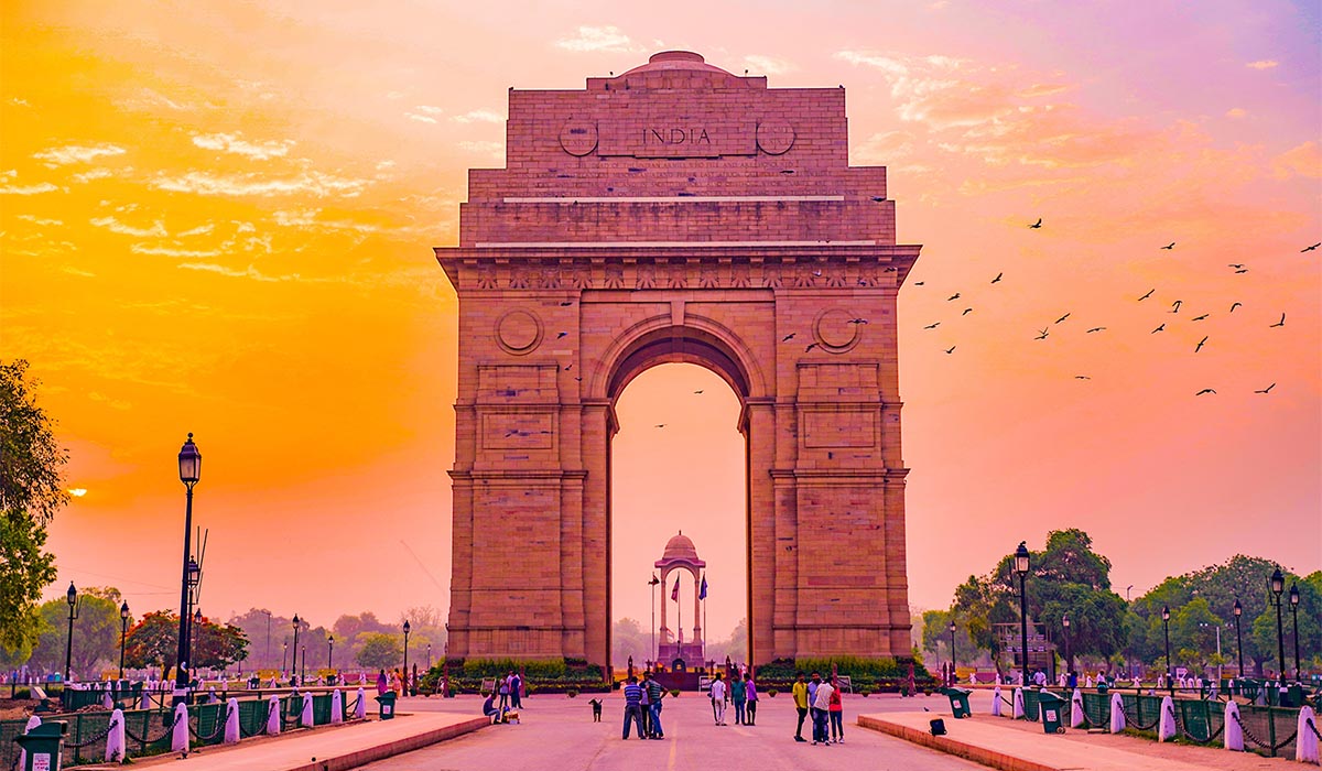 The India Gate stands tall against an orange and pink sunset. Tourists are standing beneath and taking pictures.