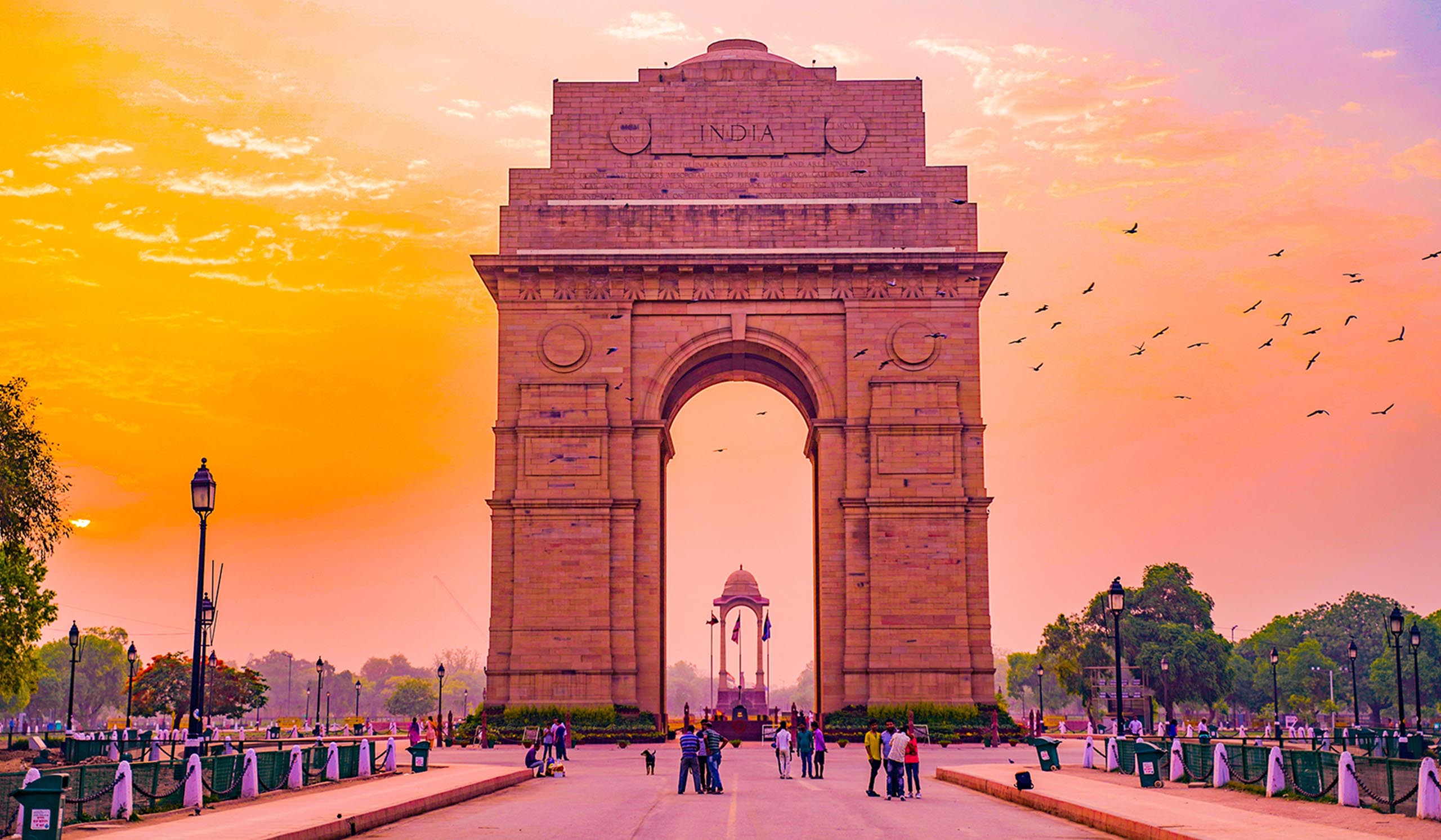 The India Gate stands tall against an orange and pink sunset. Tourists are standing beneath and taking pictures.