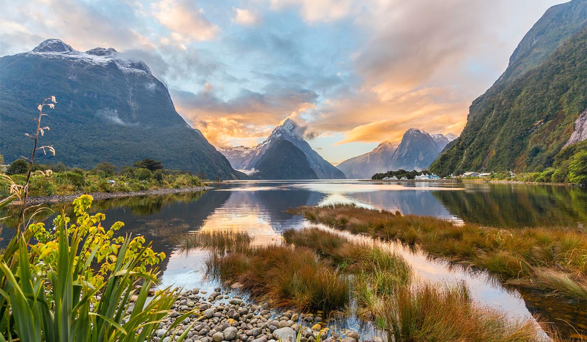 A large body of water mirrors the tall mountains and green plants surrounding it, underneath a blue, cloudy sky.