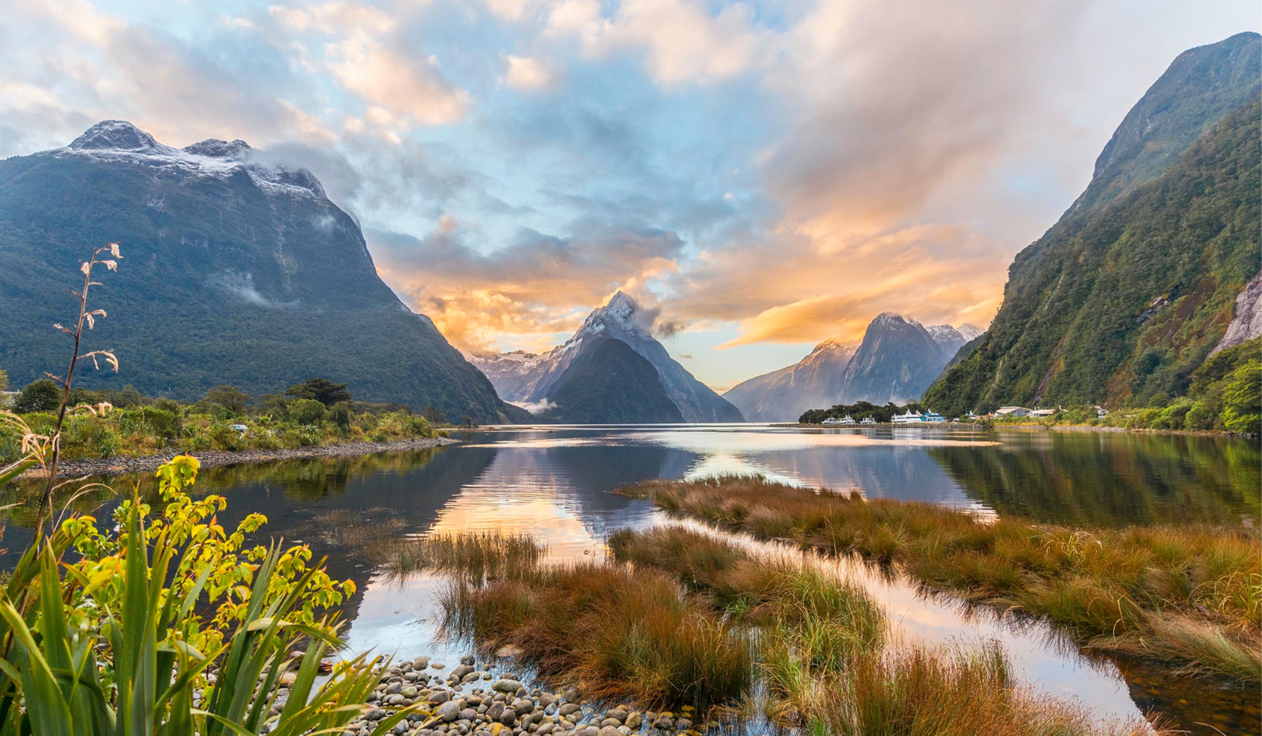 A large body of water mirrors the tall mountains and green plants surrounding it, underneath a blue, cloudy sky.