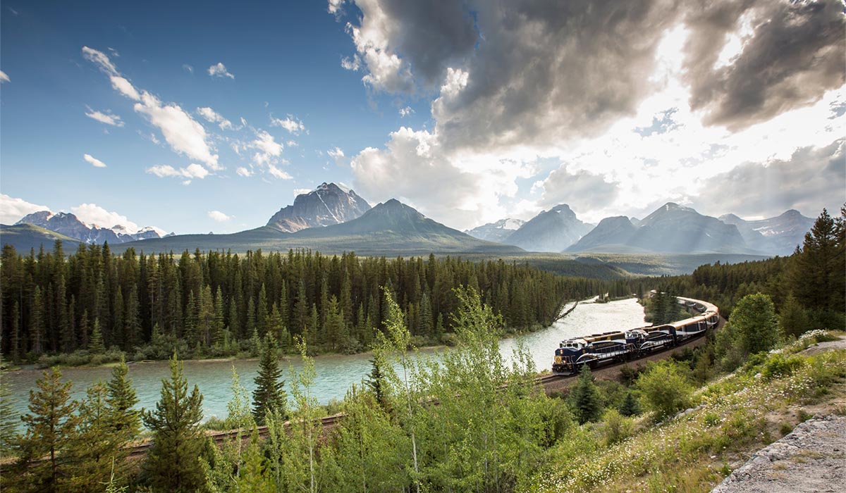 A train traveling through tall, skinny green trees along a river, with green mountains in the background under a light blue, cloudy sky.