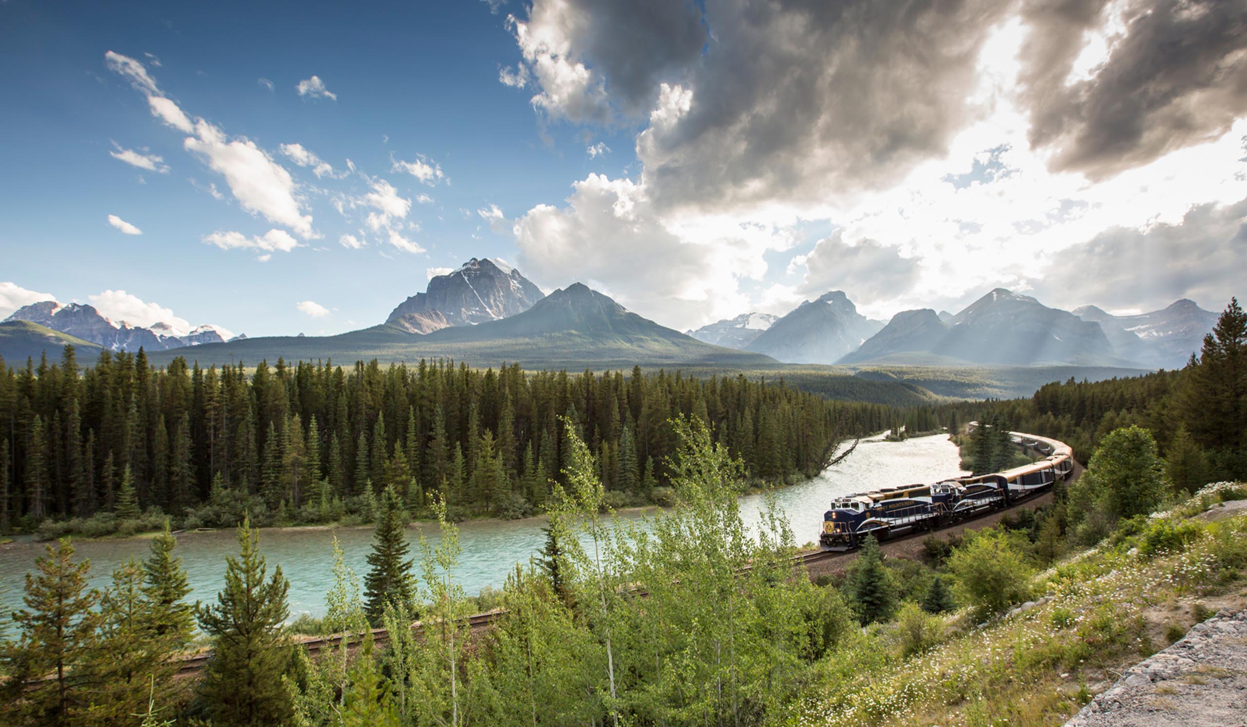 A train traveling through tall, skinny green trees along a river, with green mountains in the background under a light blue, cloudy sky.