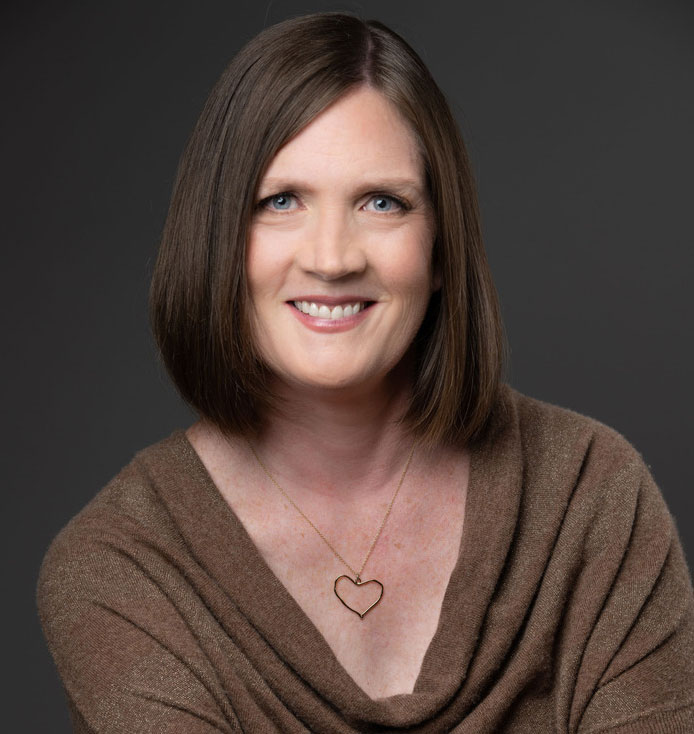 Carla Greenan, who has straight, shoulder-length brown hair and is wearing a brown top, smiles for a headshot on a dark gray background.