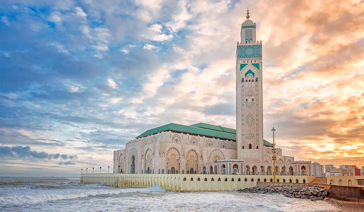 Large mosque with a green-tiled roof set along the ocean, waves crashing against the seawall at sunset under a partly cloudy sky.