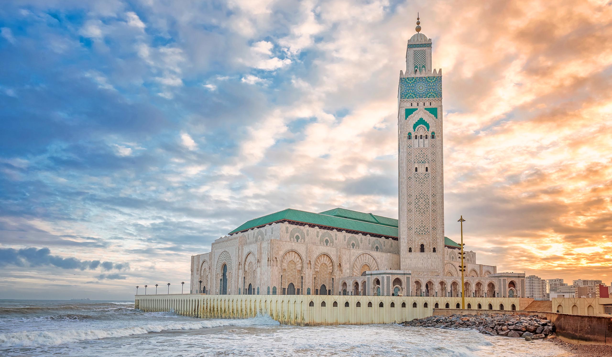 Large mosque with a green-tiled roof set along the ocean, waves crashing against the seawall at sunset under a partly cloudy sky.