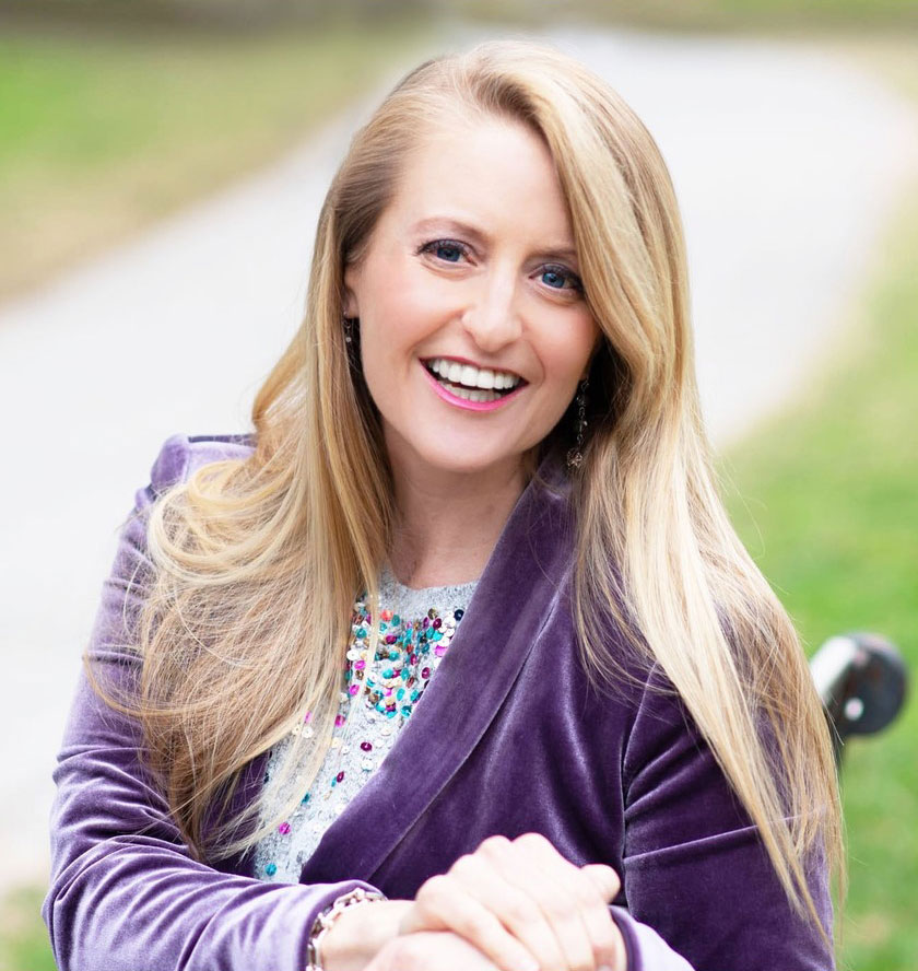 Christina Zini, who has long blonde hair and is wearing a purple velvet jacket over a sparkly, colorful top, is seated outside with blurred greenery and a pathway in the background.
