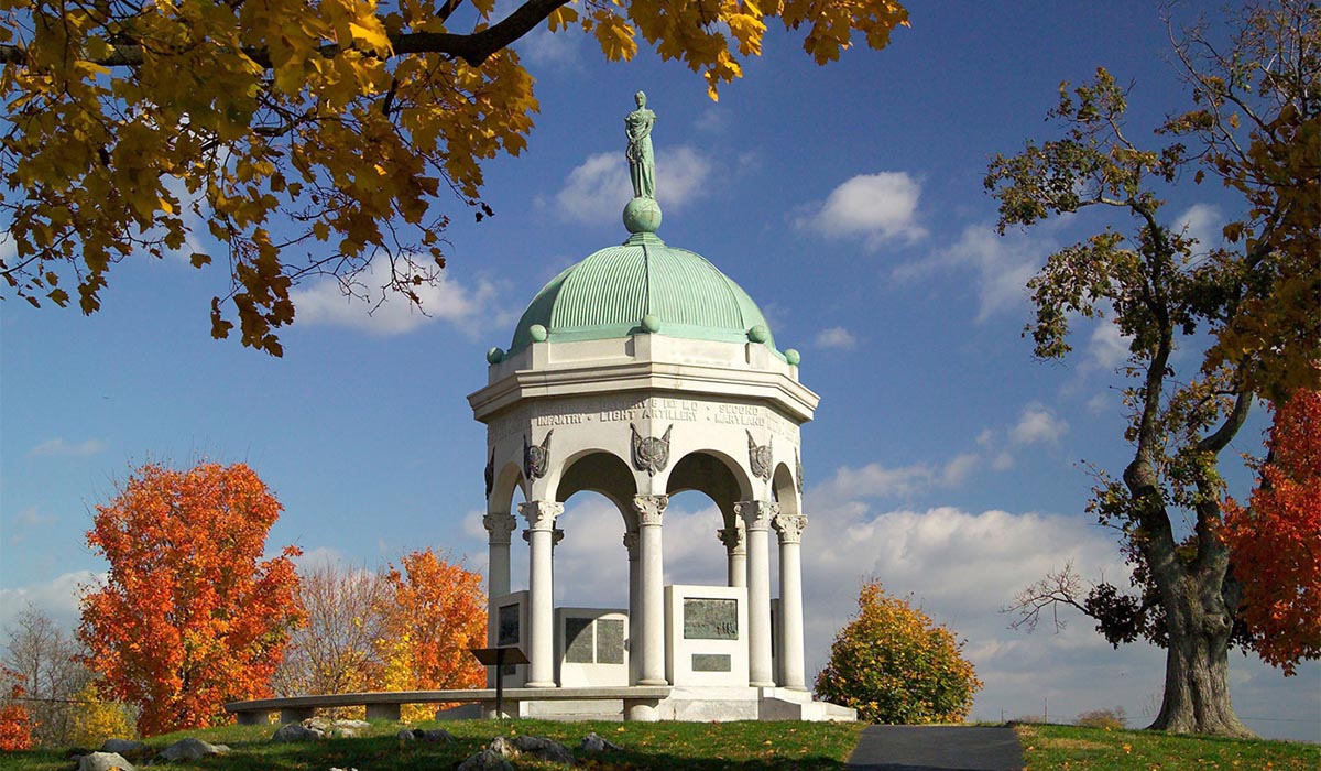 White memorial with arched columns and a green domed roof topped by a statue, surrounded by trees with bright fall foliage under a blue sky.