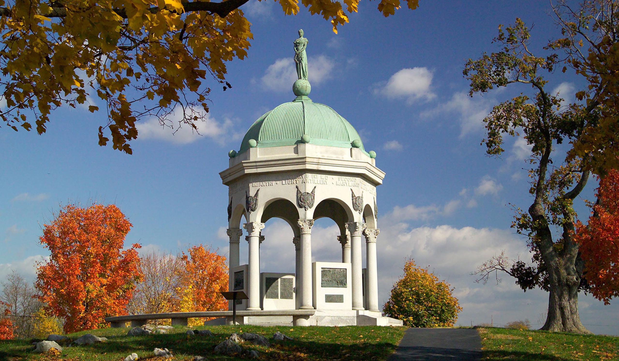 White memorial with arched columns and a green domed roof topped by a statue, surrounded by trees with bright fall foliage under a blue sky.