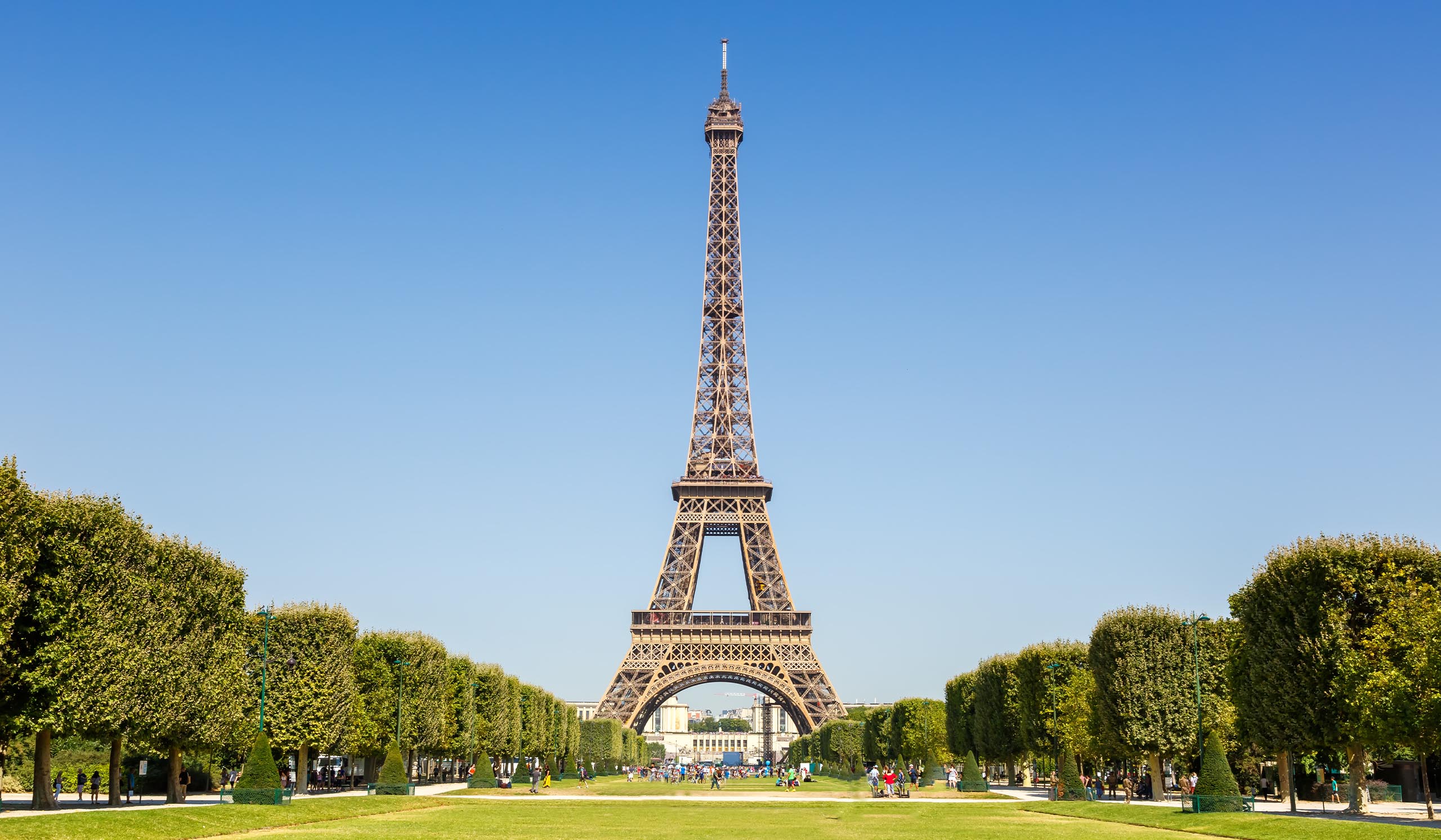 The Eiffel Tower stands against a blue sky with green trees running parallel alongside.