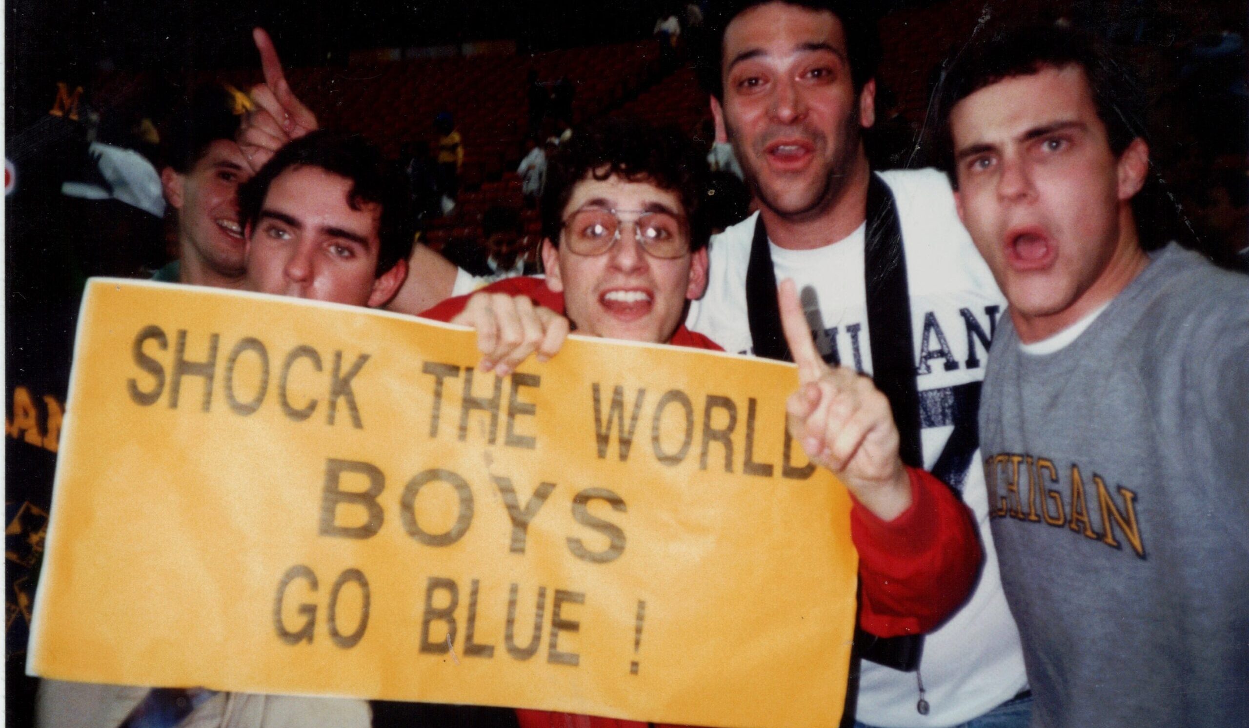 A photo from 1989, with all five friends in Michigan gear holding a yellow sign that says "Shock the world boys go blue!"