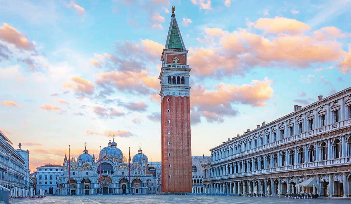 Iconic Venetian square featuring a tall brick bell tower and ornate cathedral with domes, surrounded by elegant arcaded buildings under a pastel sunset sky.