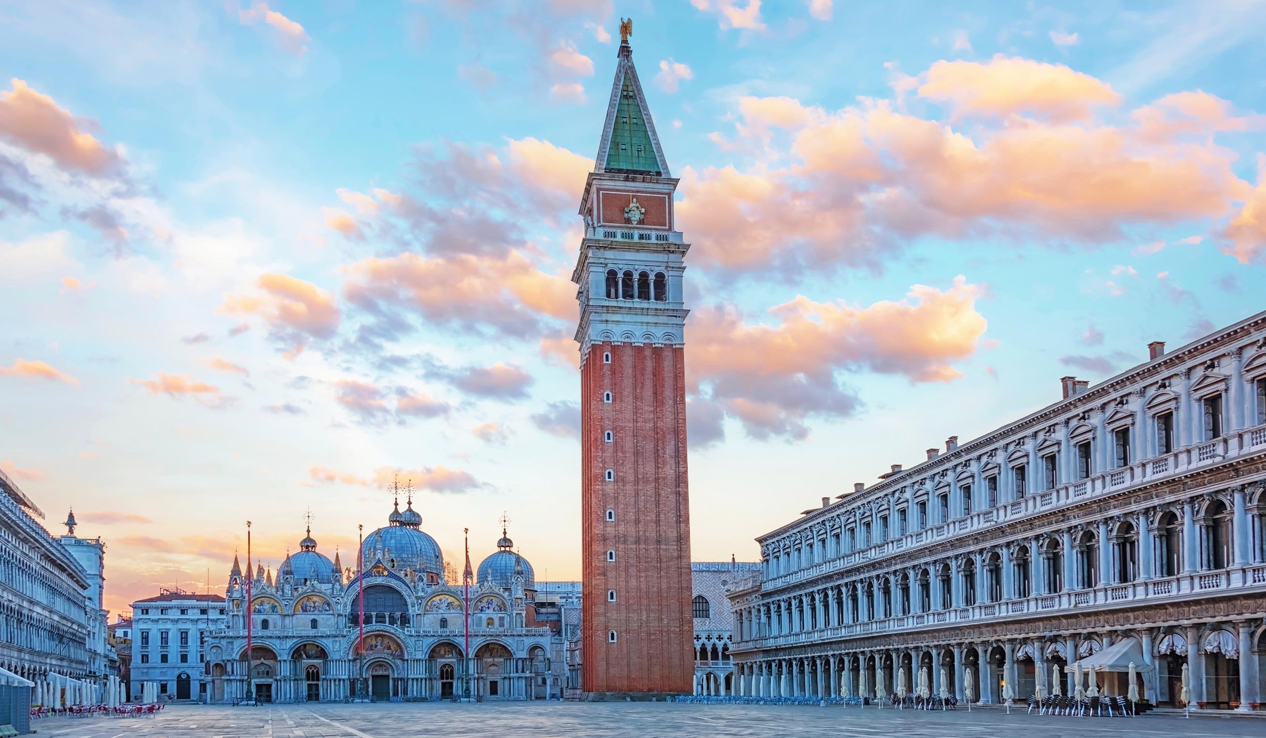 Iconic Venetian square featuring a tall brick bell tower and ornate cathedral with domes, surrounded by elegant arcaded buildings under a pastel sunset sky.