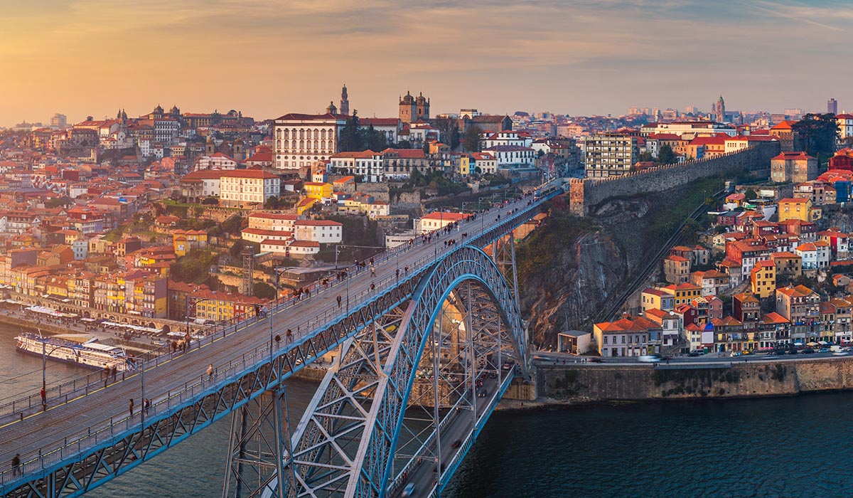 A large arch bridge spans a river, connecting a hillside city with colorful buildings under a warm sunset sky.