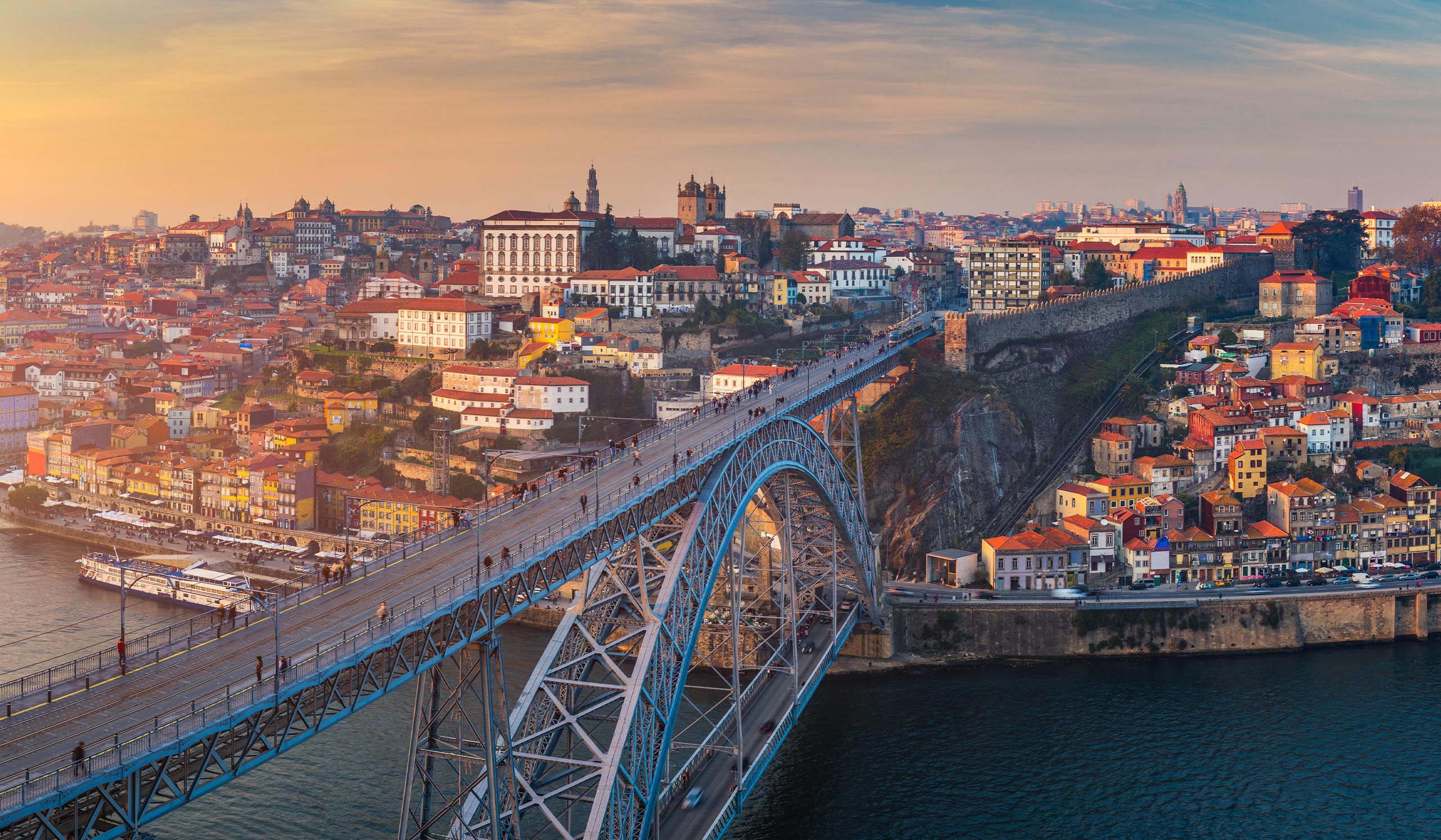 A large arch bridge spans a river, connecting a hillside city with colorful buildings under a warm sunset sky.