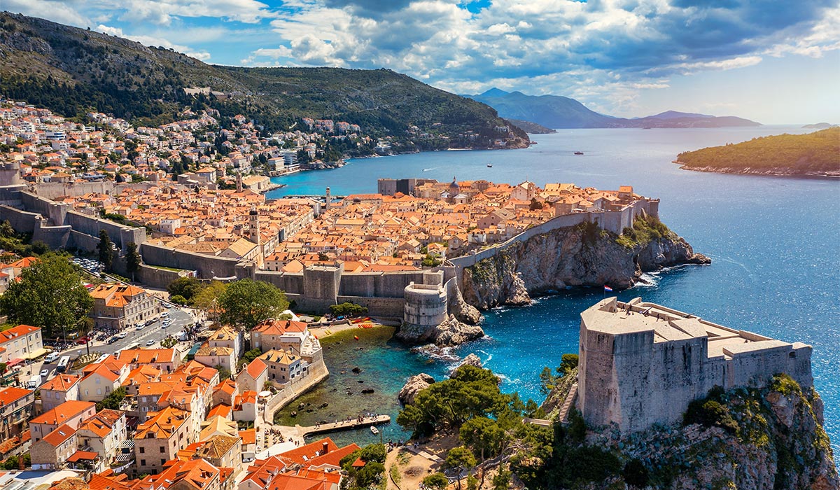 A packed town of tan buildings aligns along the coast of vibrant blue waters, and green mountains in the background.