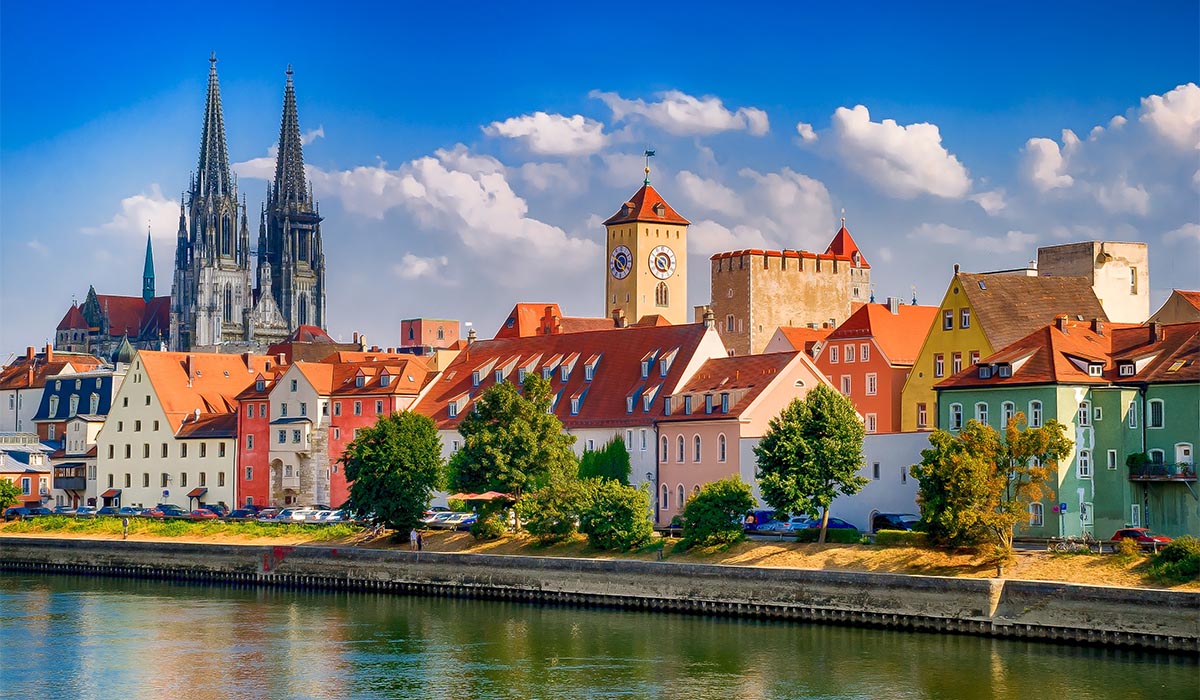 Colorful buildings line a river, with a tall cathedral rising above the city under a bright blue sky.
