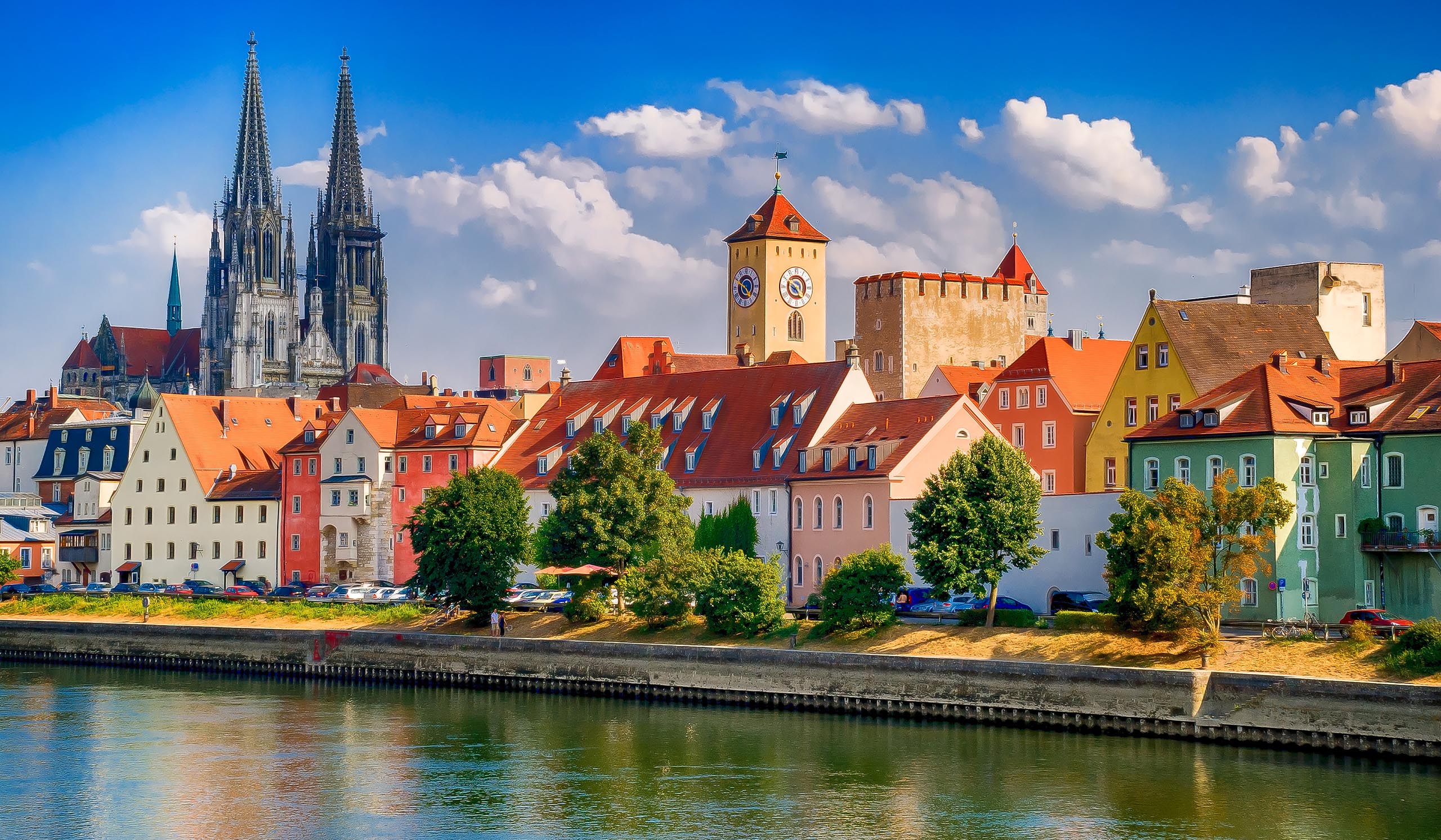 Colorful buildings line a river, with a tall cathedral rising above the city under a bright blue sky.