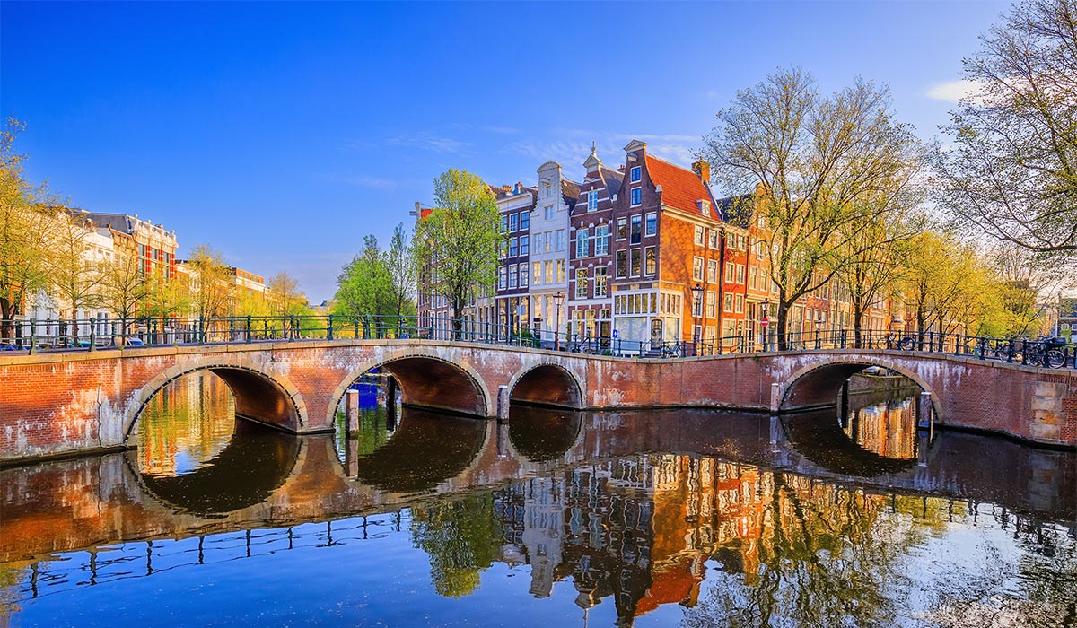 A stone bridge crosses a canal with reflections of colorful buildings and trees under a clear blue sky.