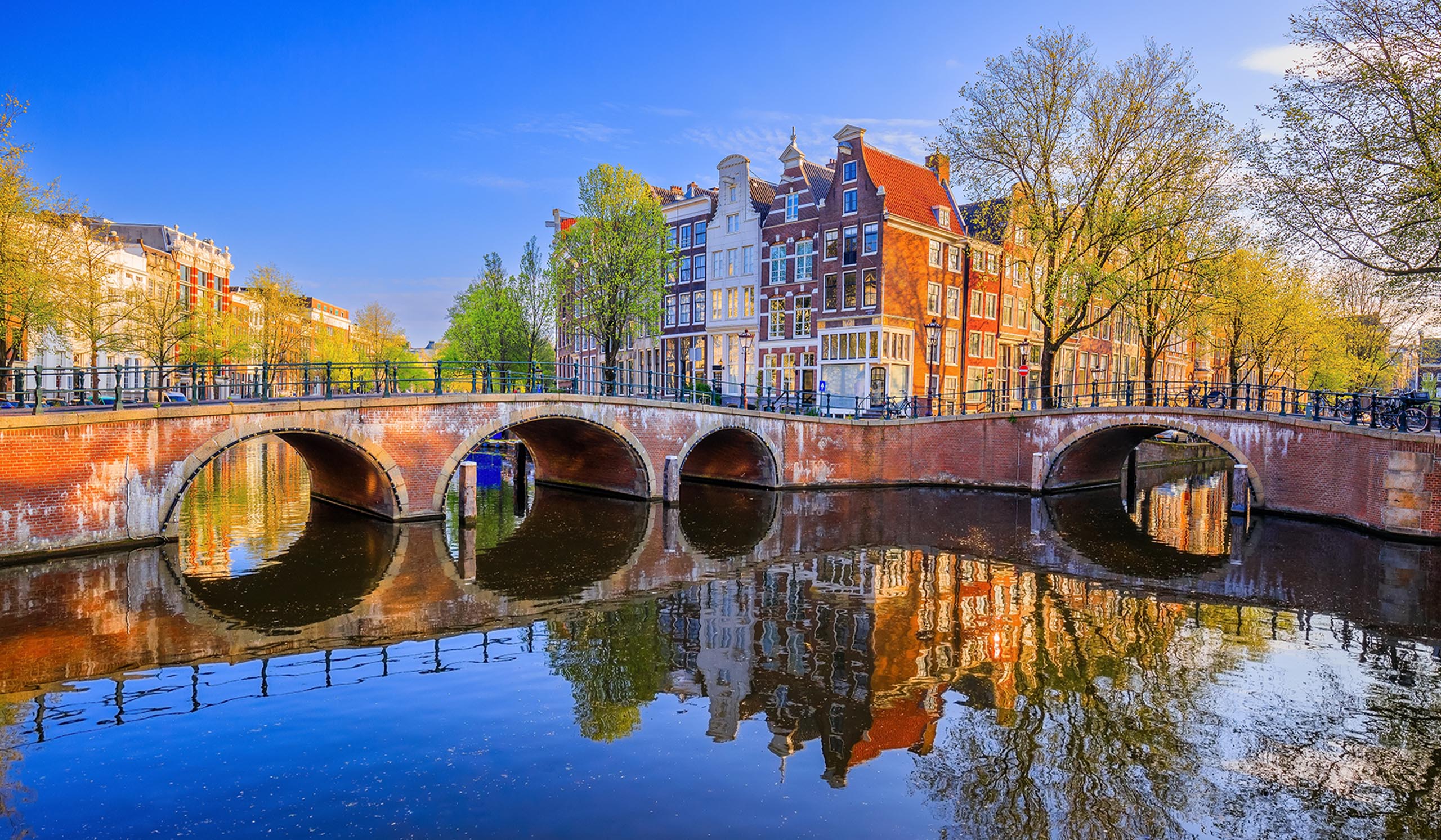 A stone bridge crosses a canal with reflections of colorful buildings and trees under a clear blue sky.
