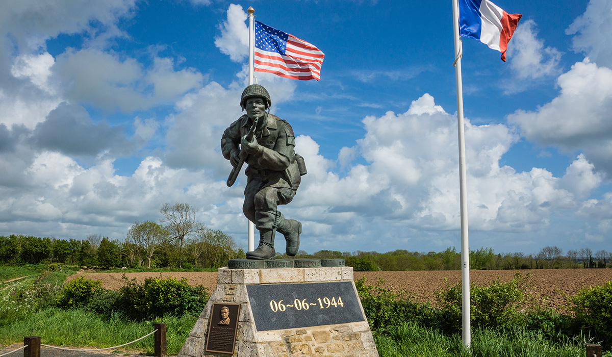 Statue of a soldier advancing with a rifle stands on a pedestal marked “06-06-1944,” with American and French flags flying behind it under a partly cloudy sky.