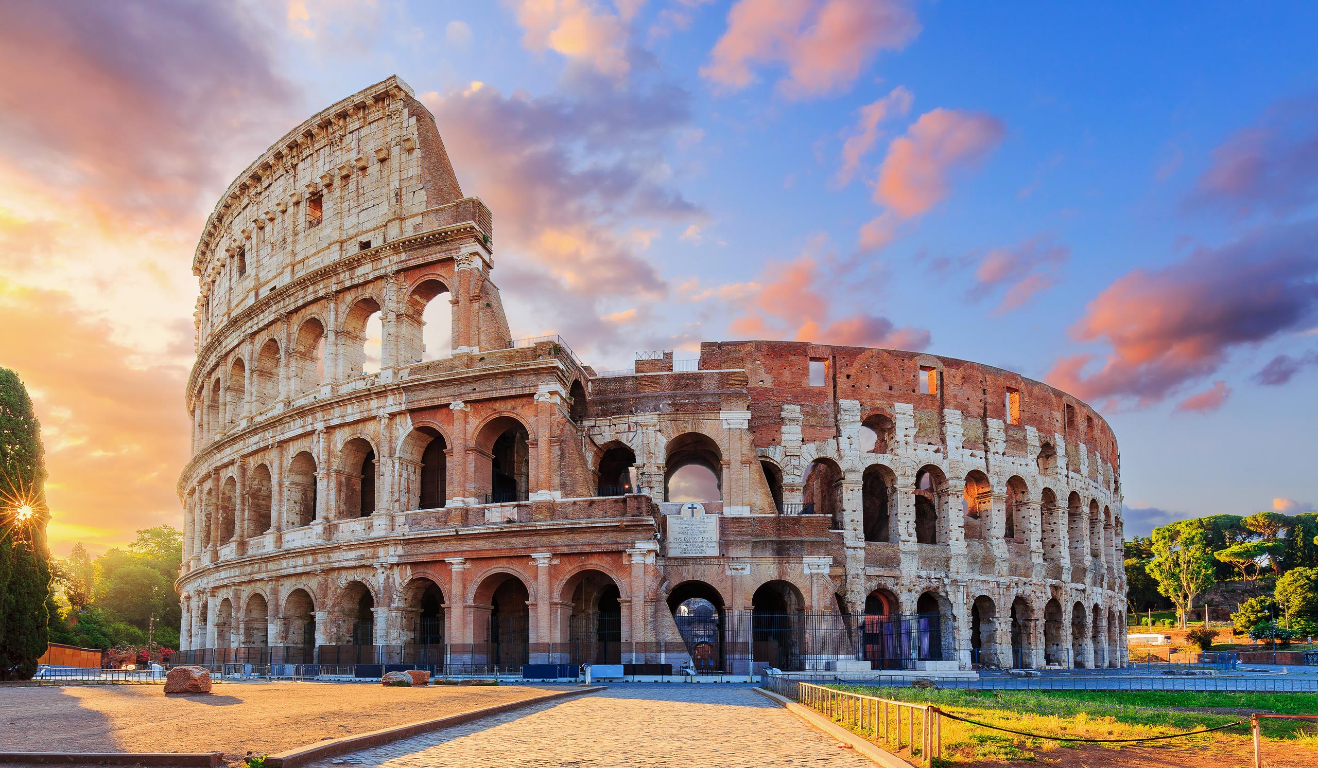 The Colosseum stands in Rome under a colorful sunset sky.