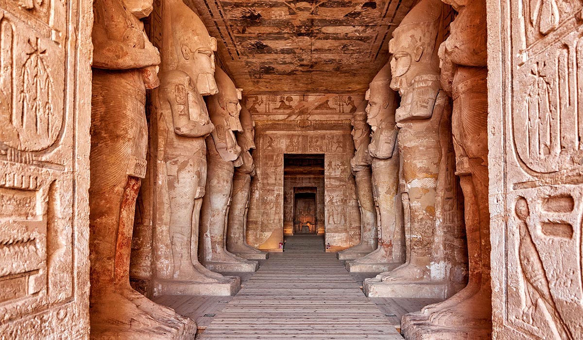 Interior corridor of an ancient Egyptian temple lined with large carved statues and hieroglyph-covered walls, leading to a doorway in the distance.